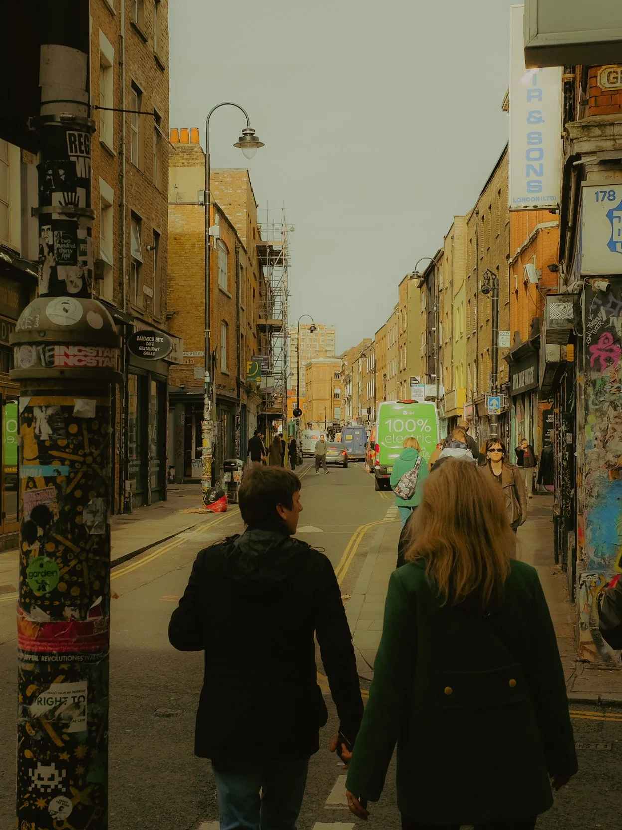 Rue urbaine animée avec des piétons et des véhicules, bâtiments en brique, lampadaires, devantures de magasins, affiches, graffiti, et un ciel nuageux.