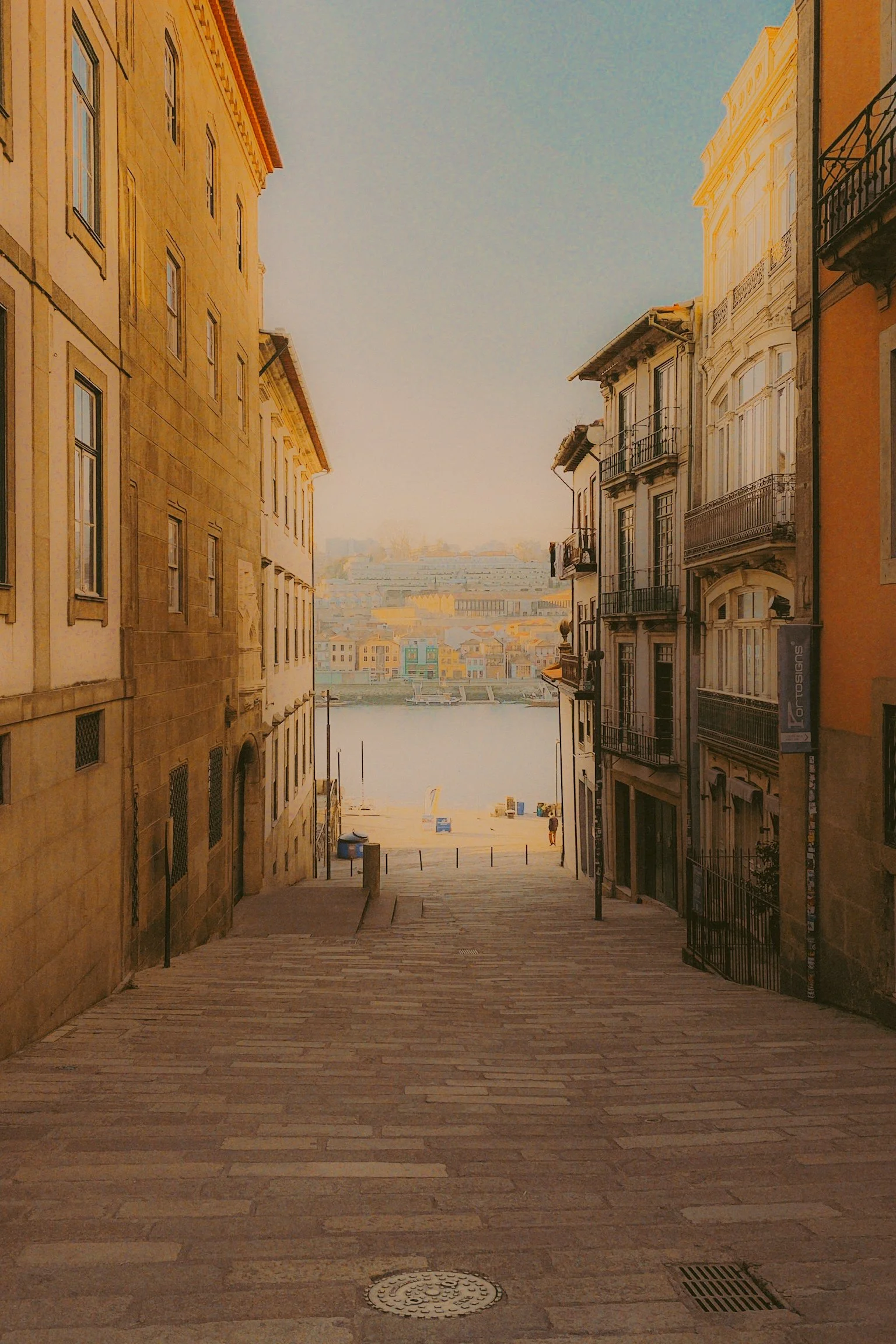Vue d'une ruelle descendante entre des immeubles anciens menant à une place près d'une rivière, avec des bâtiments colorés au loin et un ciel clair.