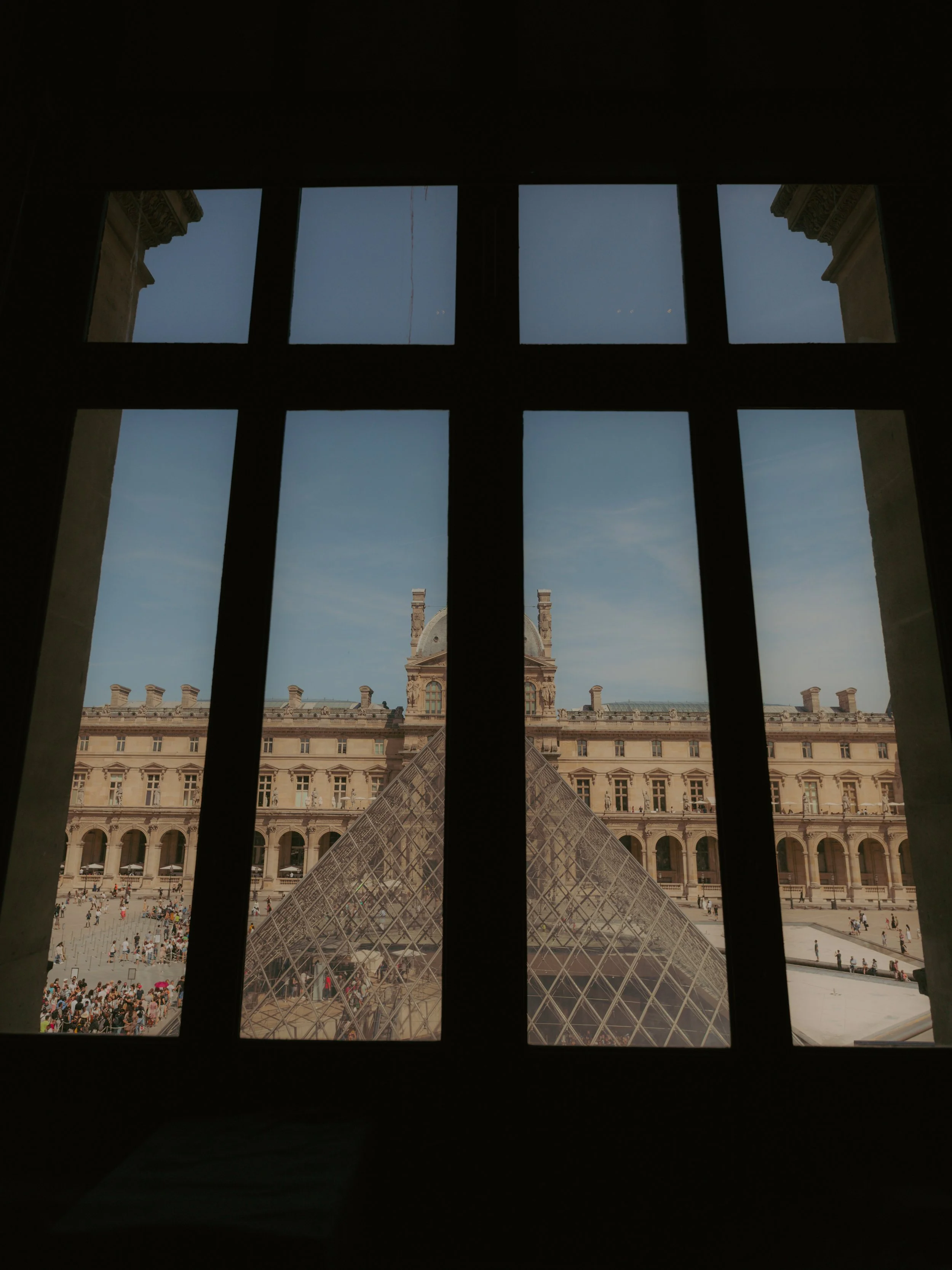 Vue de la pyramide du Louvre à travers une fenêtre avec volets, avec un bâtiment historique en arrière-plan et un ciel bleu clair.