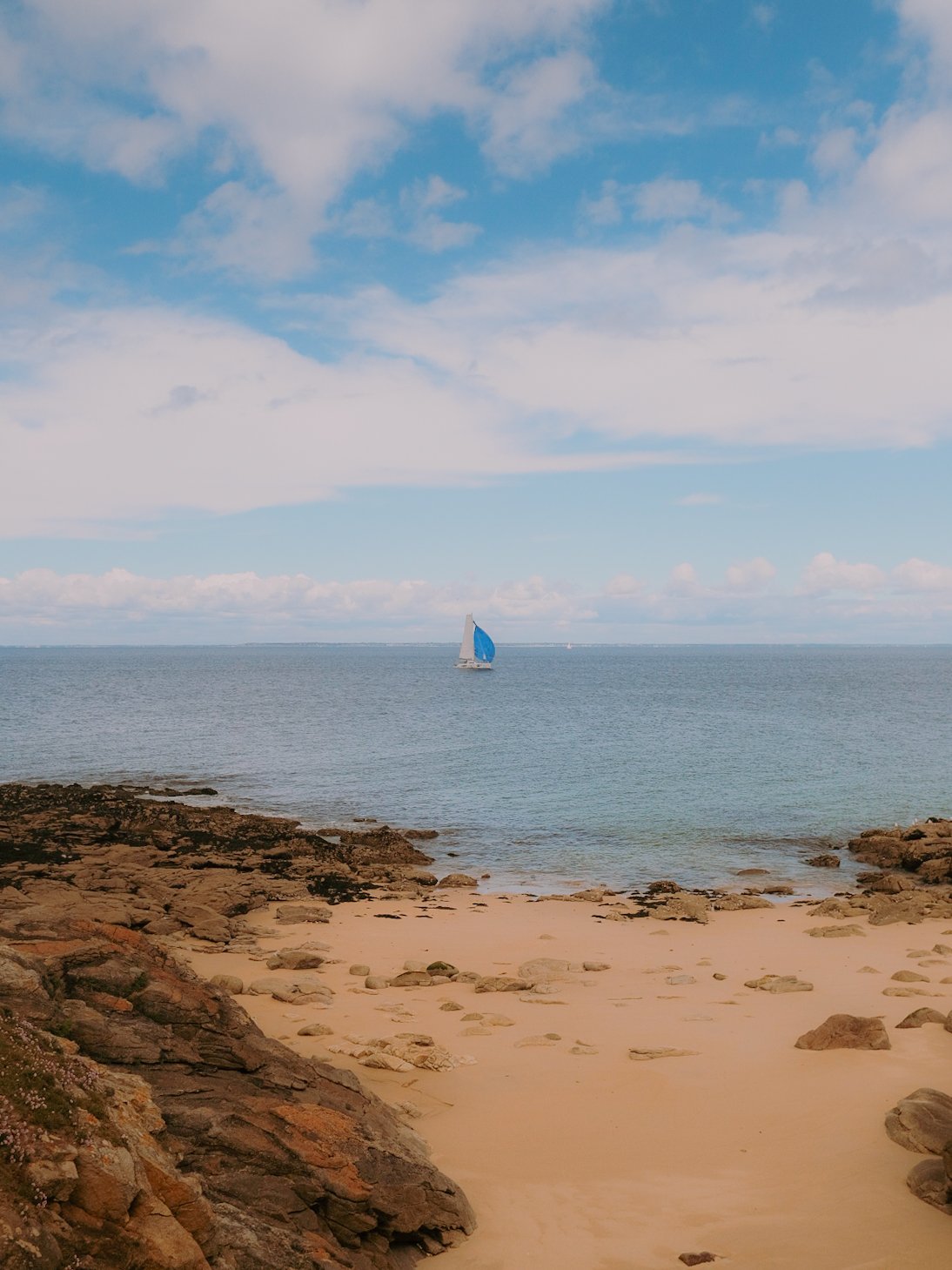 Plage rocheuse avec sable, océan et voilier à l'horizon sous un ciel partiellement nuageux.