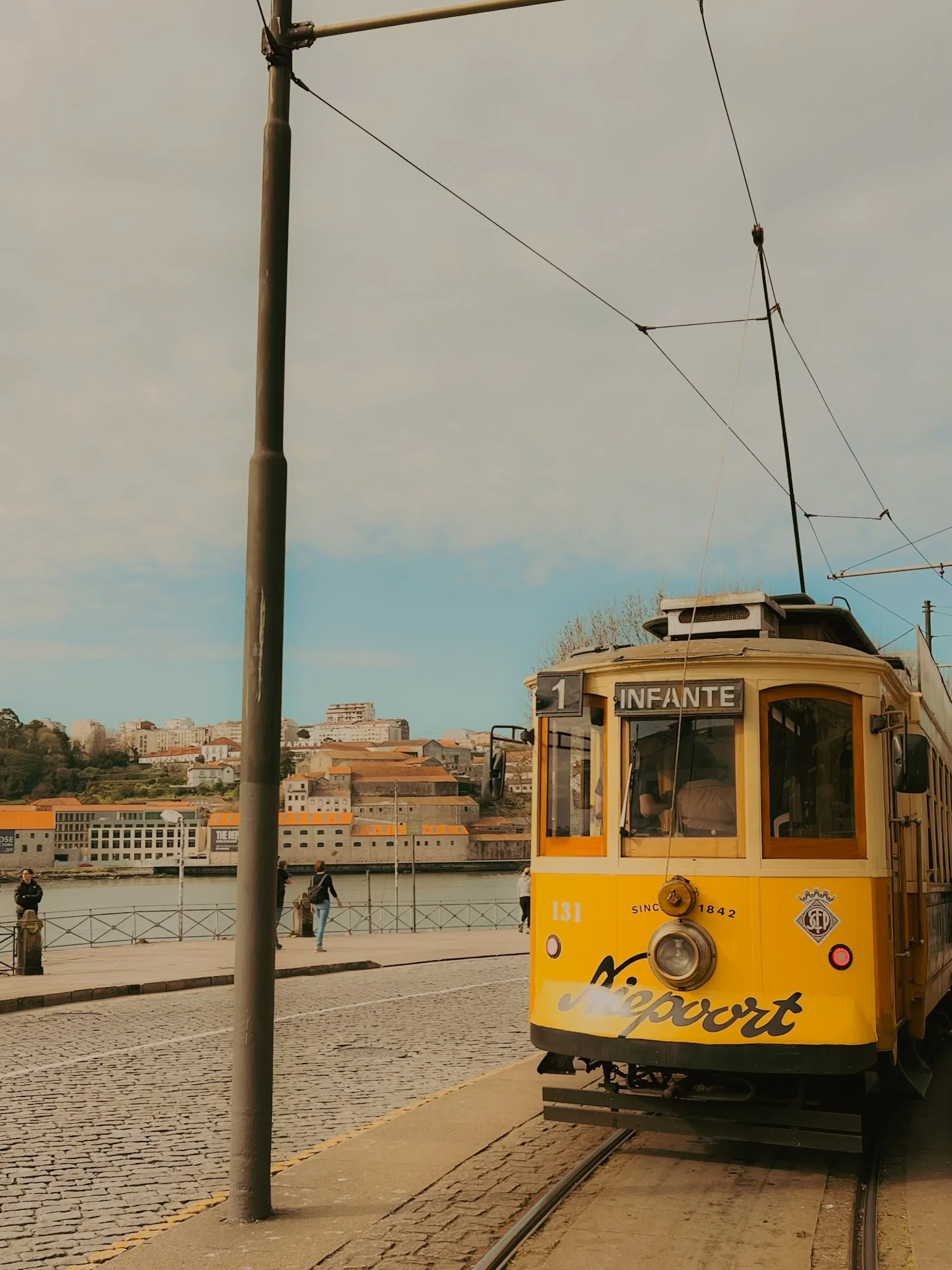 Un tram jaune avec le mot "Naveport" écrit dessus, indiquant la ligne 1 avec la destination "Infante". Il circule près d'une rivière ou d'un fleuve, avec une ville en arrière-plan, sous un ciel partiellement nuageux.