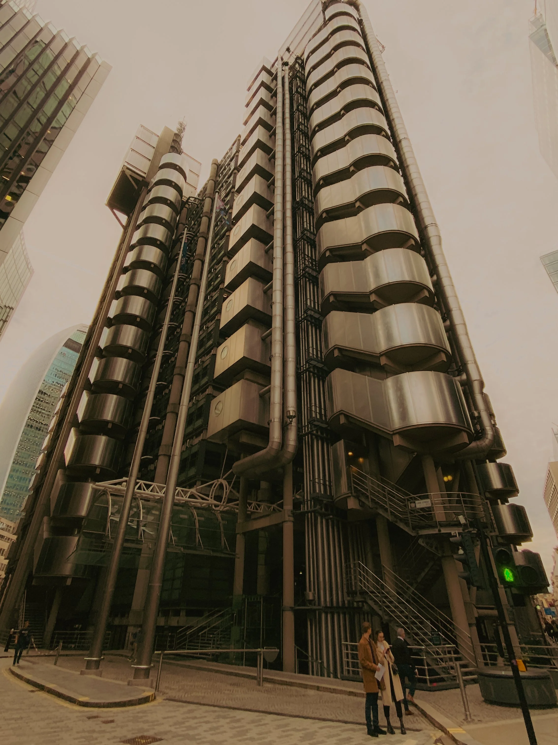 Gratte-ciel moderne avec structure métallique distinctive, grande entrée avec escalier et groupe de personnes attendant au pied du bâtiment.