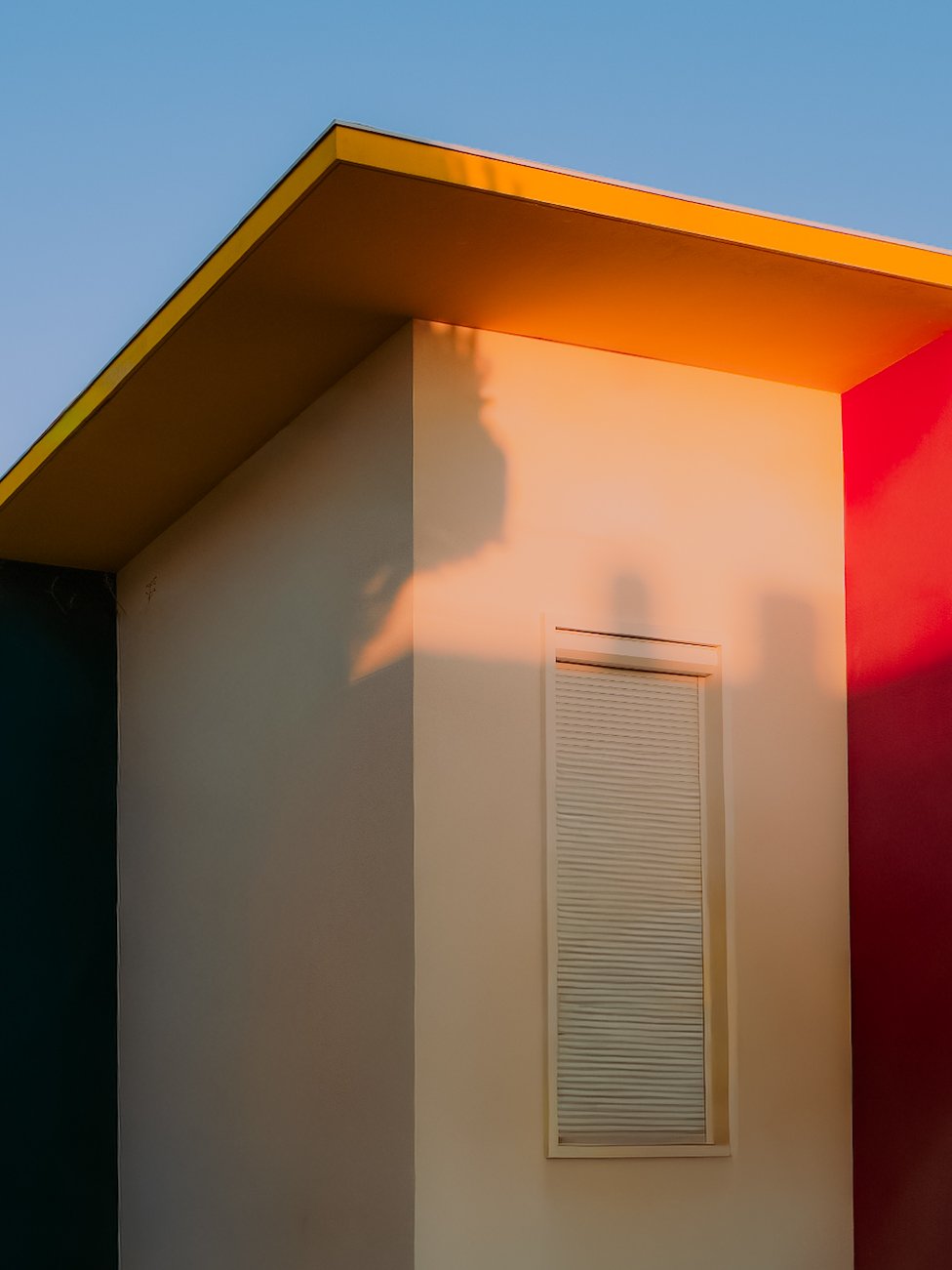 Fassade d'une maison moderne avec un mur blanc, une fenêtre avec volet roulant et un toit en pente jaune-orange, éclairée par la lumière du coucher de soleil.