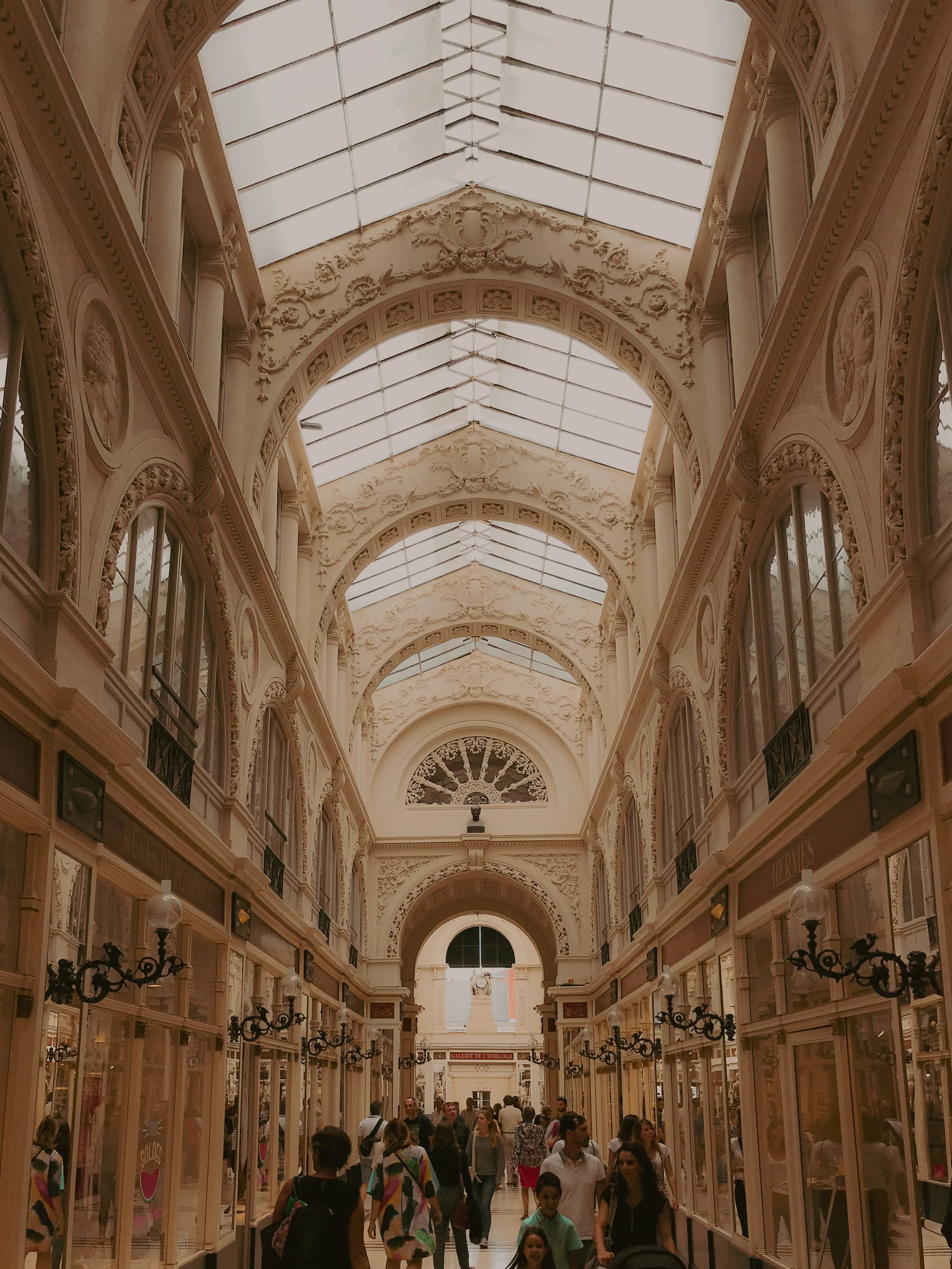Intérieur d'un grand centre commercial avec un plafond en verre orné de décorations architecturales, occupé par des nombreuses personnes.