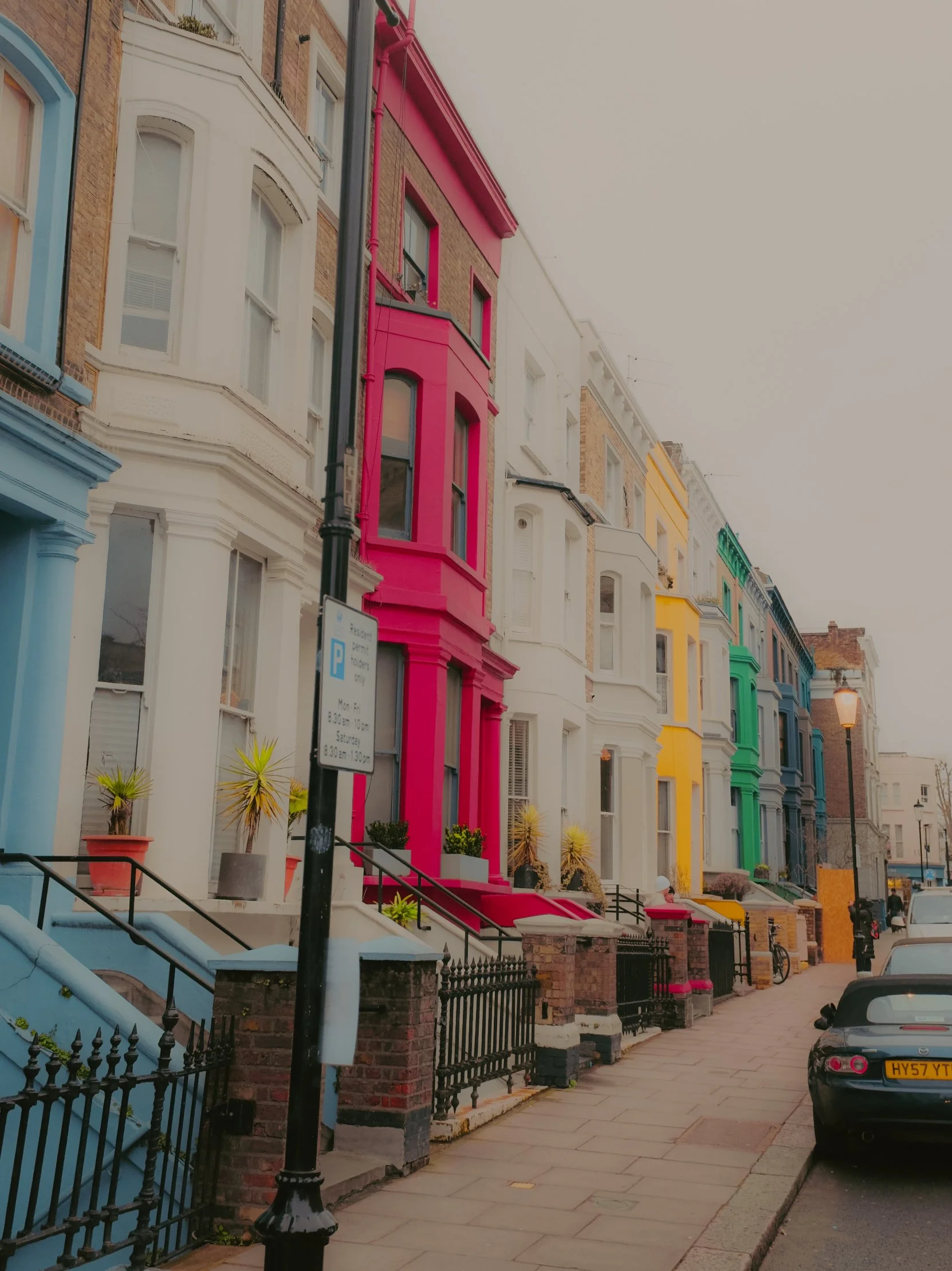 Façades colorées de maisons victoriennes avec escaliers en pierre, sur une rue pavée, quelques voitures garées et lampadaires, sous un ciel nuageux.