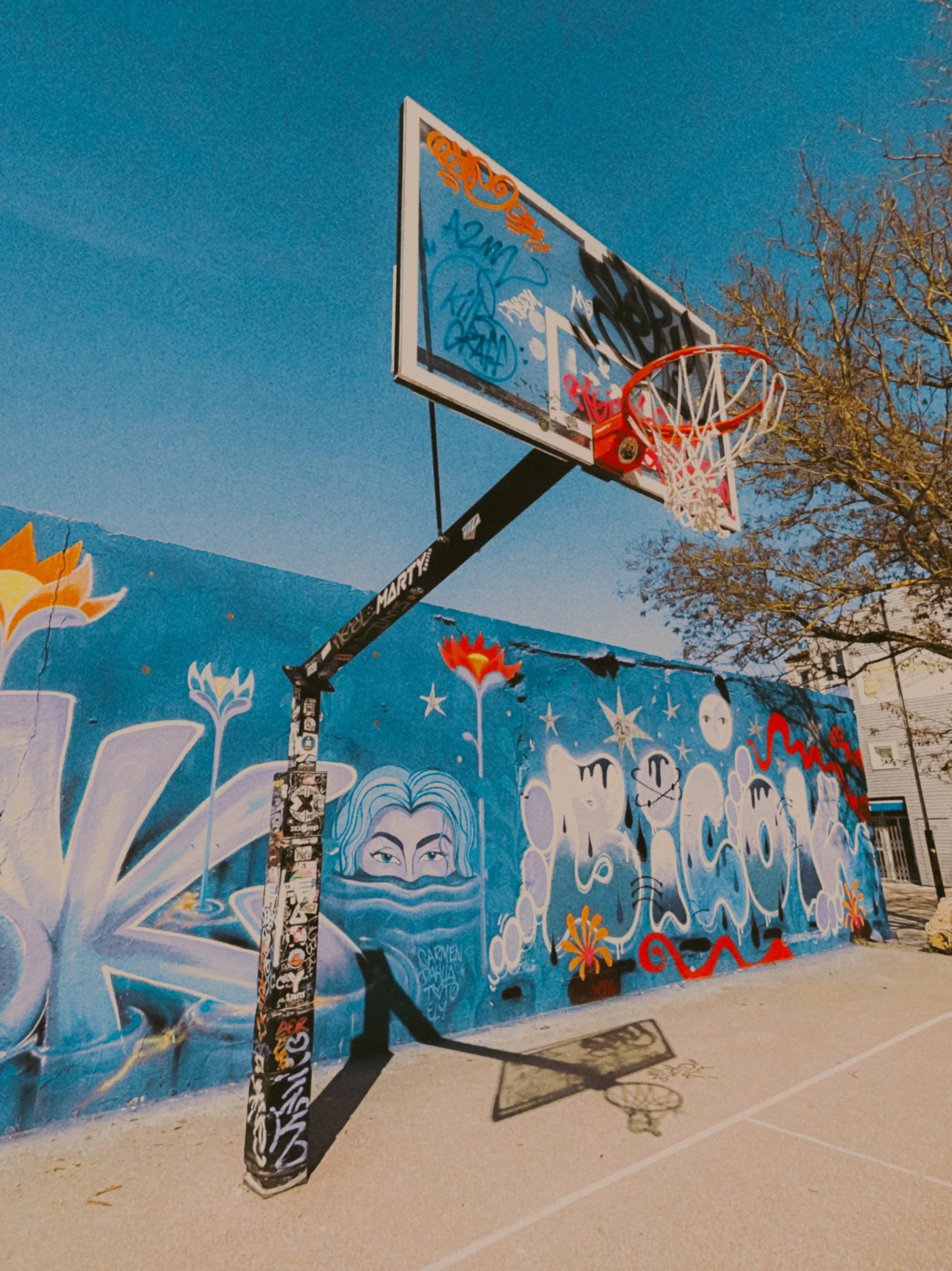 Mur de graffitis avec un tableau de basketball. Le mur est décoré de plusieurs graffitis colorés, y compris un visage de femme avec des cheveux blancs et un masque facial, plusieurs mots et dessins abstraits.