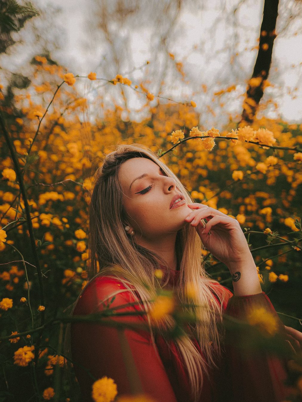 Jeune femme aux cheveux longs, portant un haut rouge, entourée de fleurs jaunes dans un jardin ou un parc.
