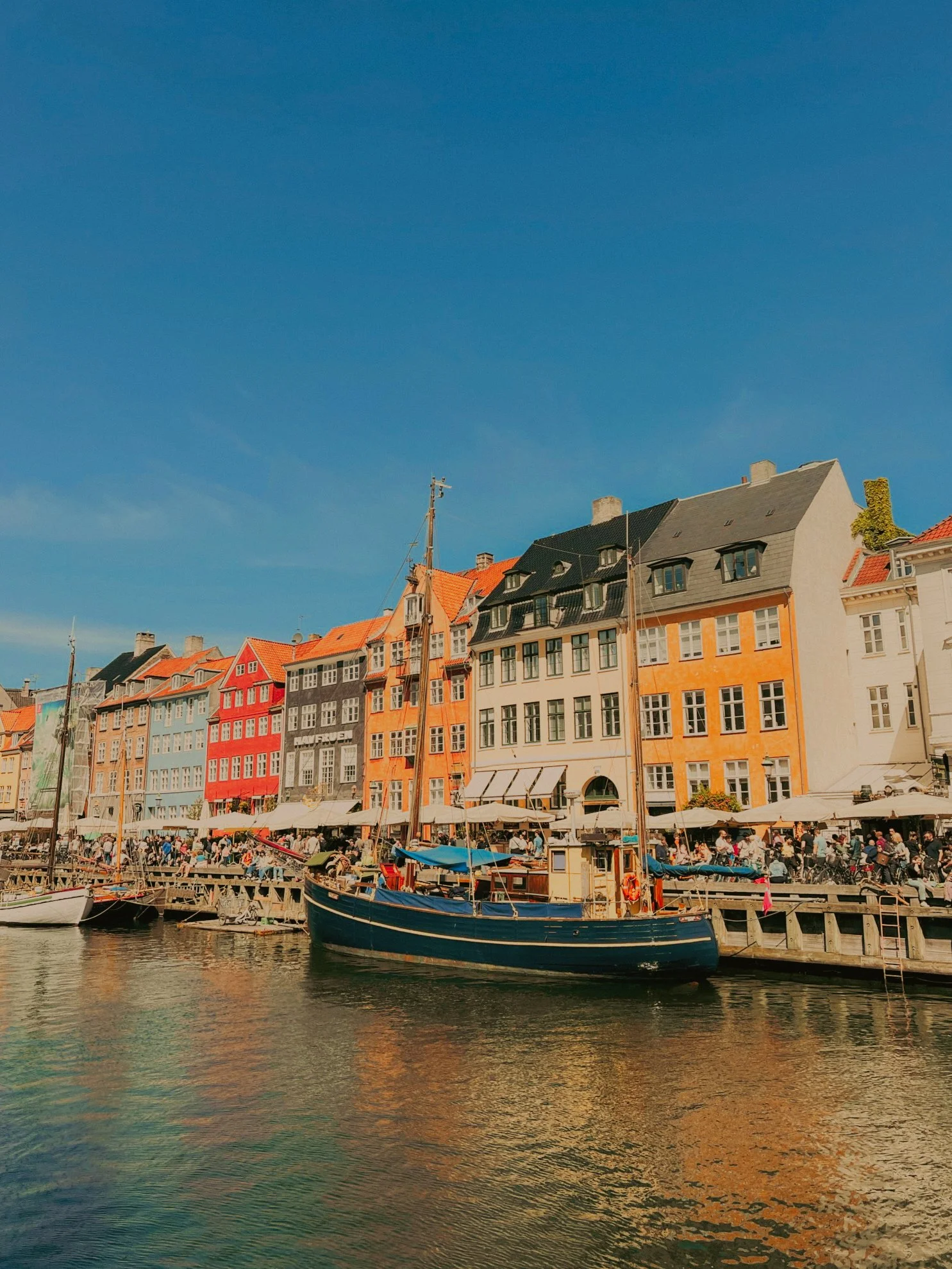Un quai avec des voiliers et des bateaux, des bâtiments colorés à l'arrière-plan, un ciel bleu, et une foule de personnes au bord de l'eau.