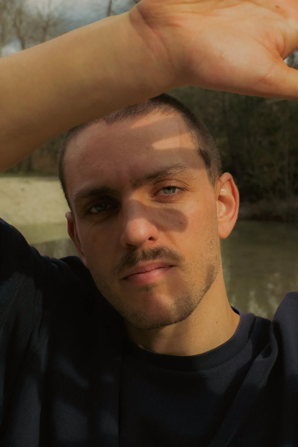 Un jeune homme aux cheveux courts et à la barbe légère, avec un regard bleu, pose à l'extérieur près d'une rivière, avec des arbres en arrière-plan. Il porte un t-shirt noir et un bras levé, laissant de la lumière du soleil et des ombres jouer sur so