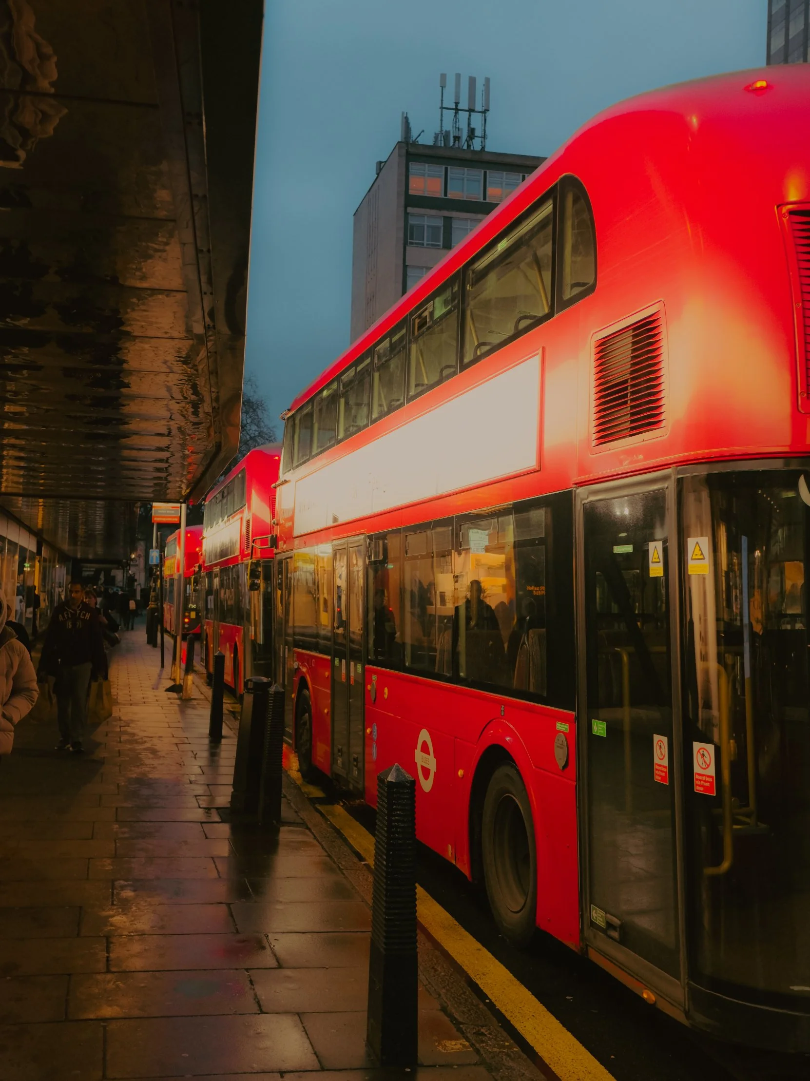 Bus à deux étages rouge stationné sur un trottoir urbain lors du crépuscule, avec des passants marchant à proximité et un bâtiment en arrière-plan.