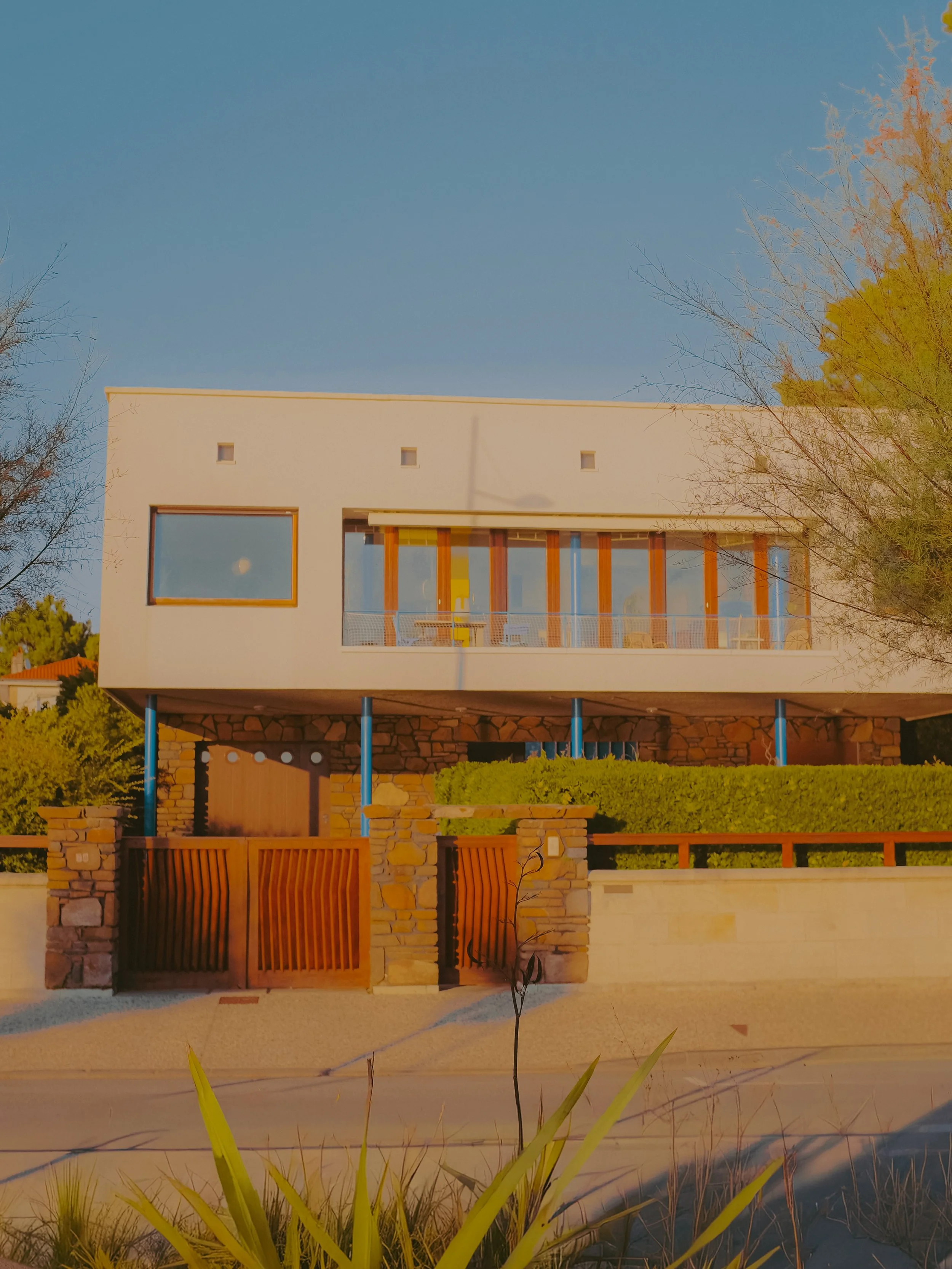 Maison moderne à deux étages avec façade blanche, grandes fenêtres, clôture en pierre et bois, arbres et ciel clair.