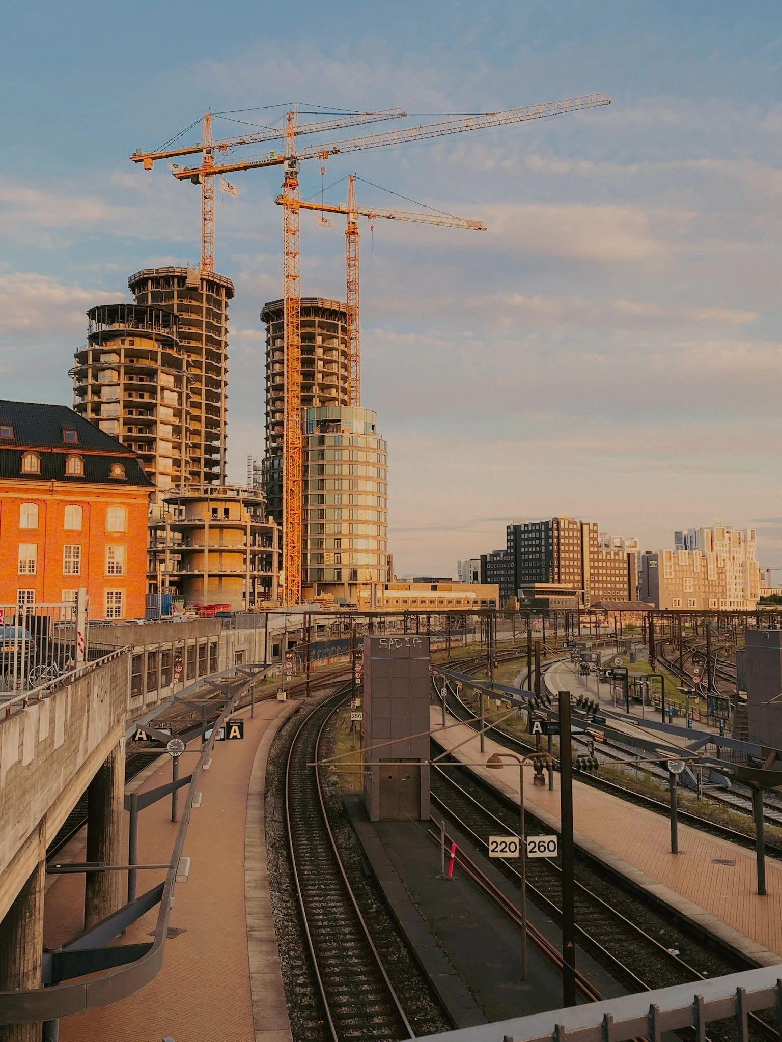 Construction de gratte-ciel avec deux grues sur un site urbain, train et rails dans un stationnement à proximité.