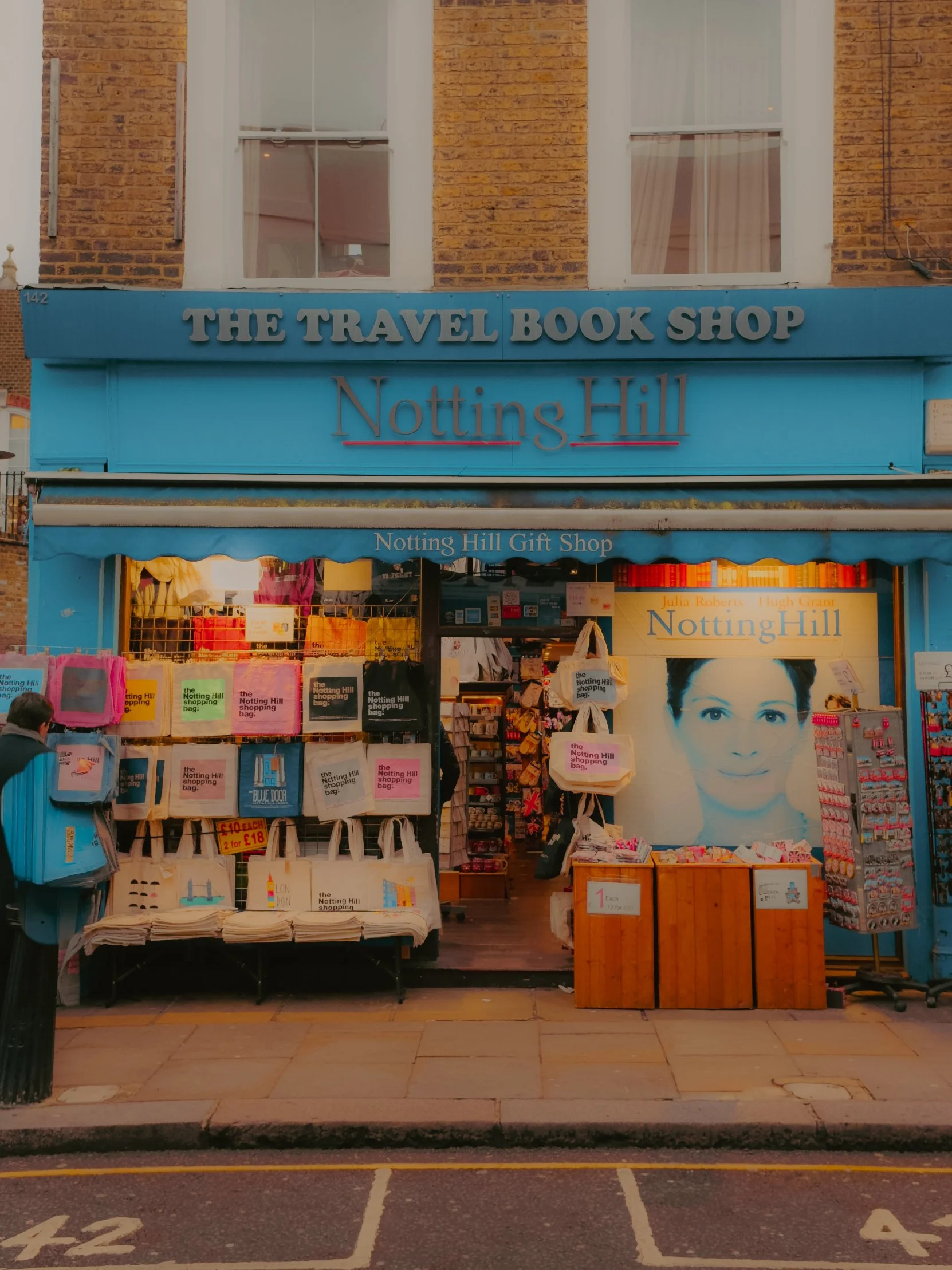 Une boutique nommée 'Nottings Hill' qui vend des livres de voyage, des sacs, des souvenirs et des cartes postales, située sur une rue avec un trottoir en pierre.