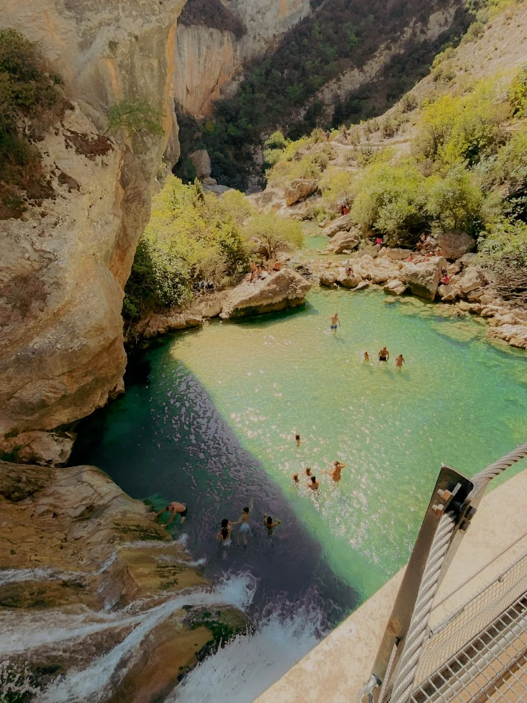 Vue d'une piscine naturelle entourée de rochers et de végétation, avec des personnes nageant et se baignant dans l'eau claire, en plein air dans une vallée montagneuse.