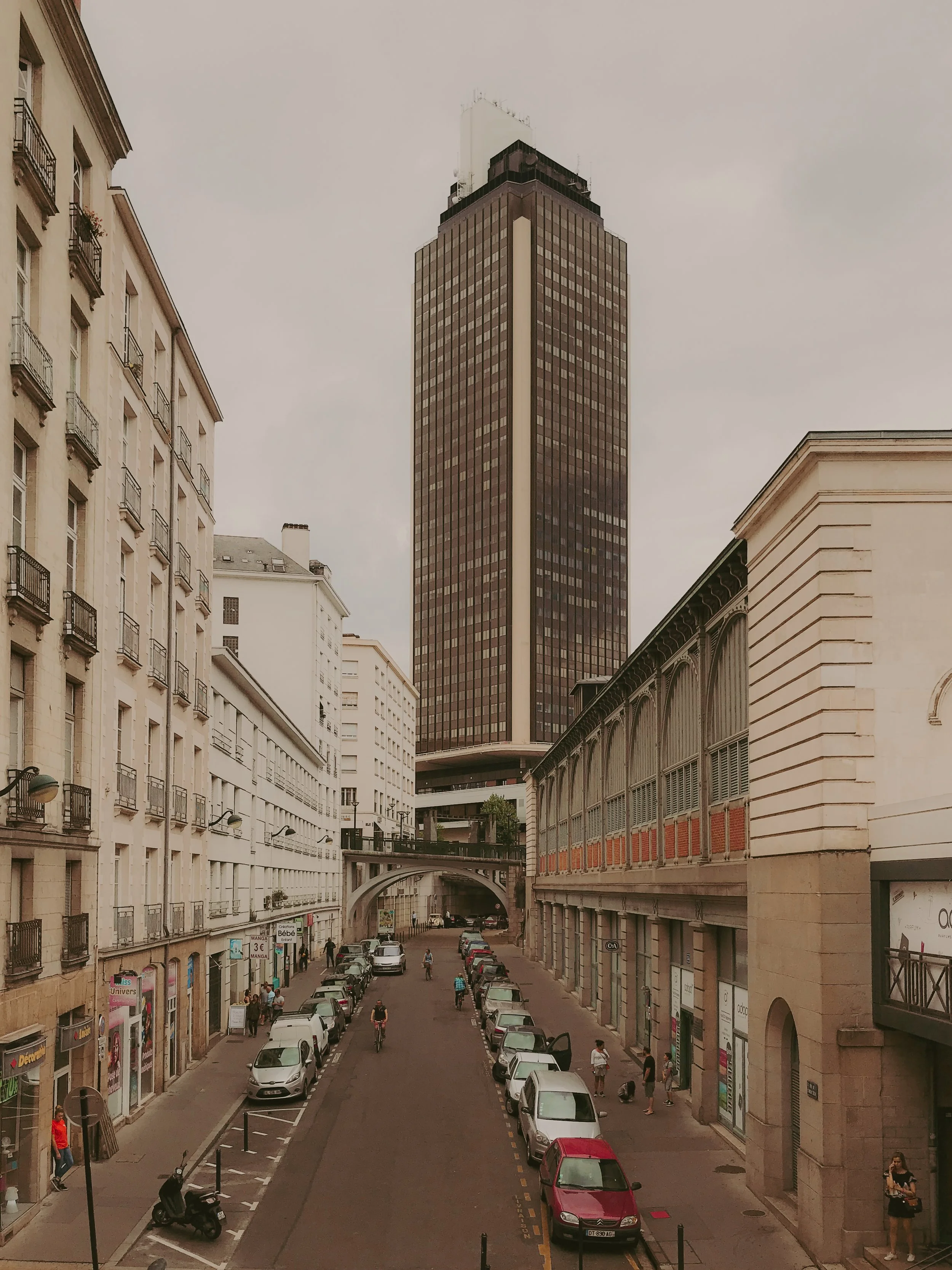 Une rue urbaine avec des voitures garées, des bâtiments résidentiels et commerciaux et une grande tour en arrière-plan, un ciel nuageux.