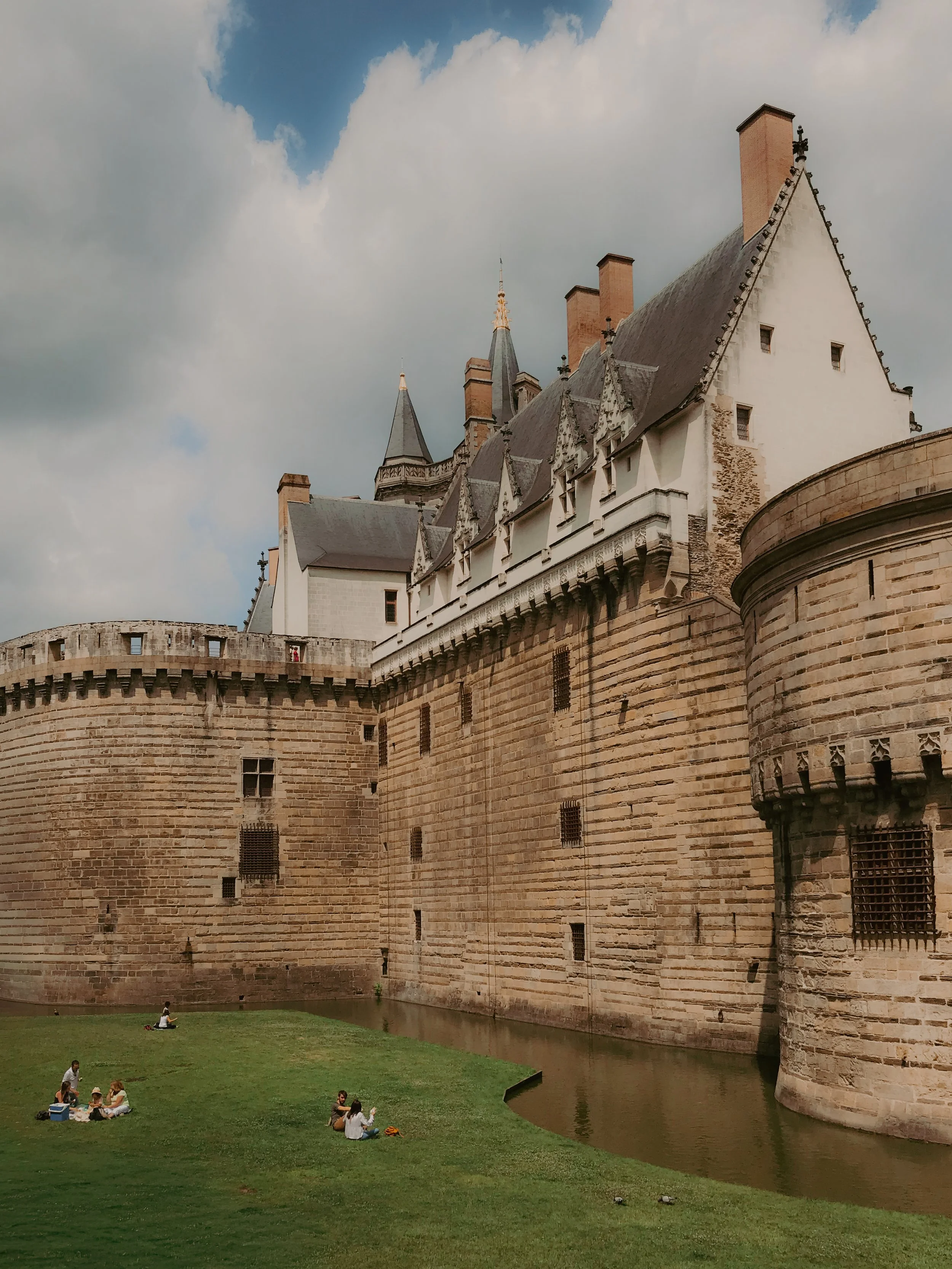 Un château médiéval avec des murs en pierre, des tours pointues et un toit en ardoise, entouré d'une faible rivière et d'une pelouse verte, avec des personnes assises sur l'herbe.