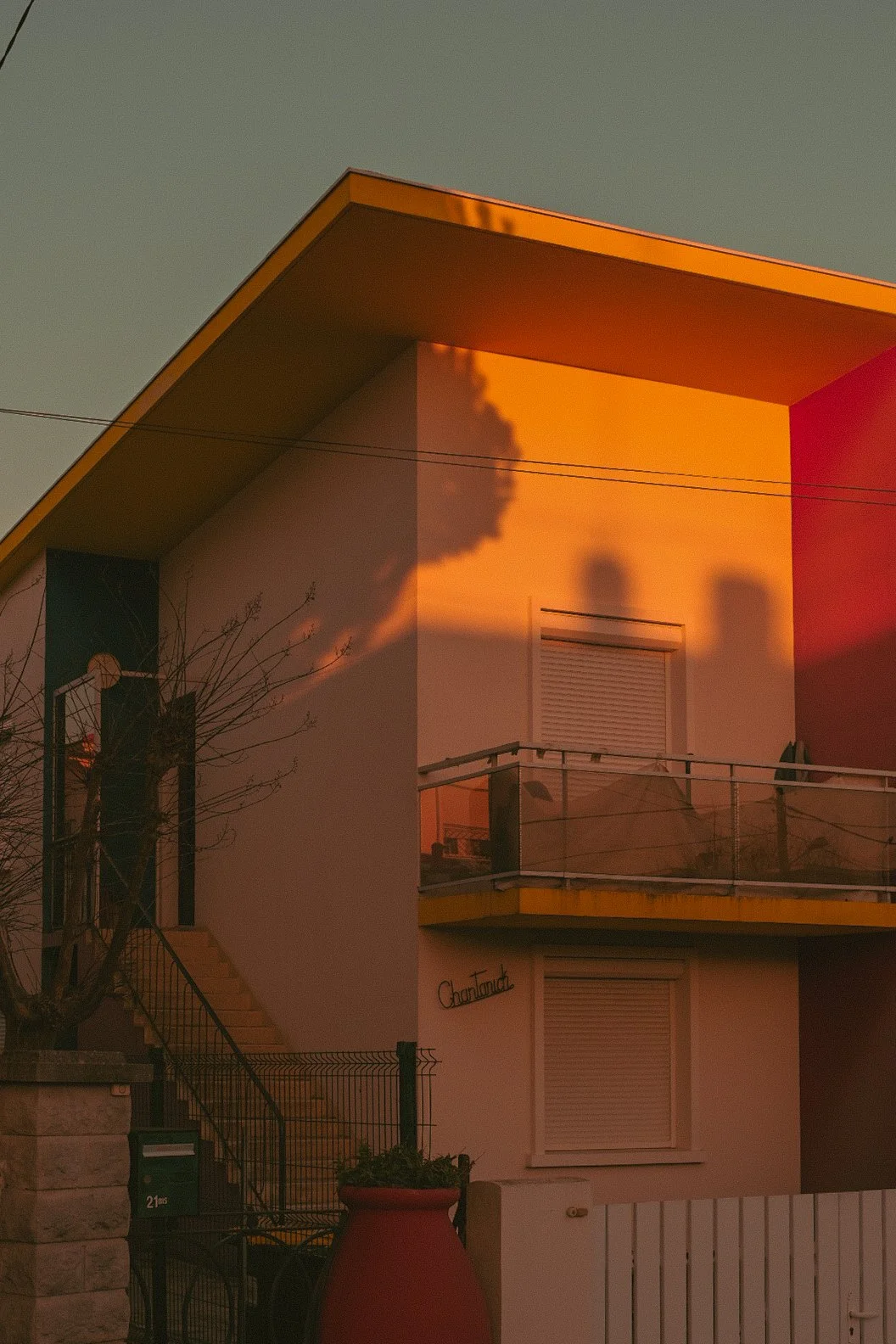 Une maison moderne avec un toit en angle, baignée par la lumière orangée du coucher de soleil, avec une balançoire sur le balcon et un arbre à proximité.