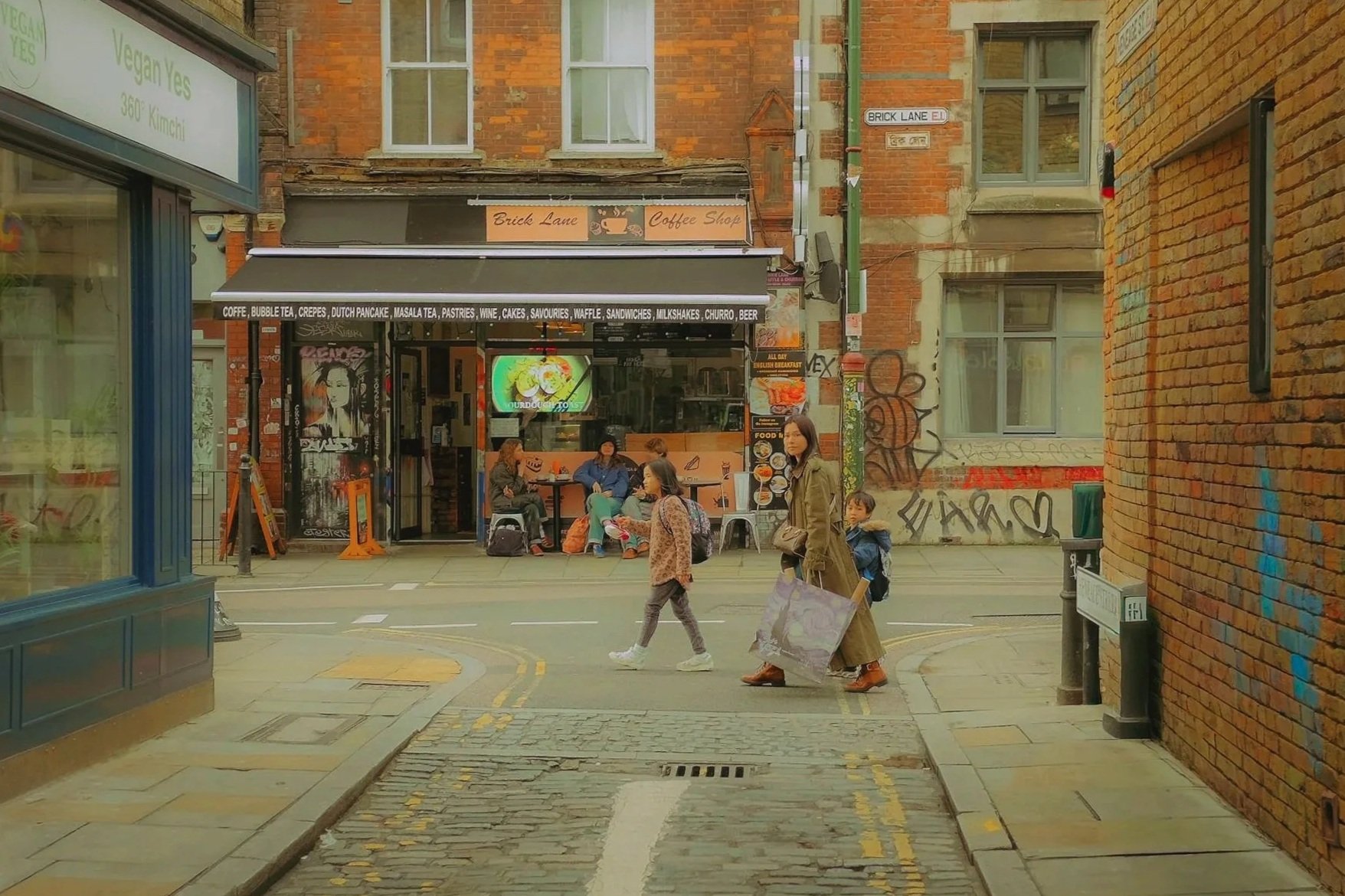 Une rue urbaine avec un café nommé 'Brick Lane Coffee Shop' en arrière-plan. Des personnes, dont une femme avec un sac en plastique, traversent la rue, et il y a des graffitis sur la mairie en arrière-plan.