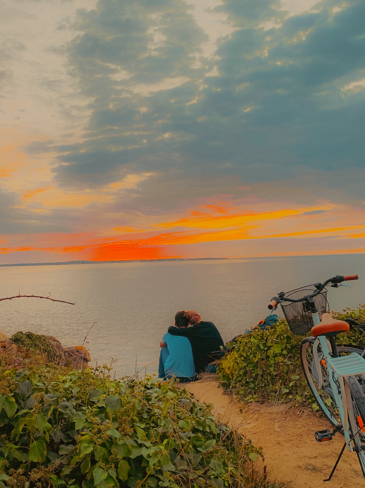 Deux personnes assises côte à côte regardant un coucher de soleil sur une étendue d'eau, avec un vélo à proximité sur un sentier entouré de végétation.