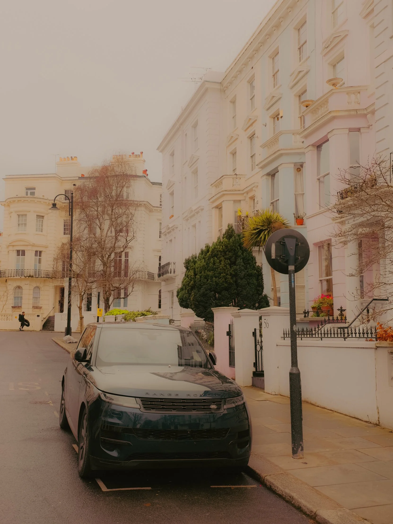 Vue d'une rue résidentielle avec des bâtiments en pierre blanche, une voiture noire devant un trottoir, des arbres sans feuilles, et un lampadaire. Le ciel est gris et brumeux.