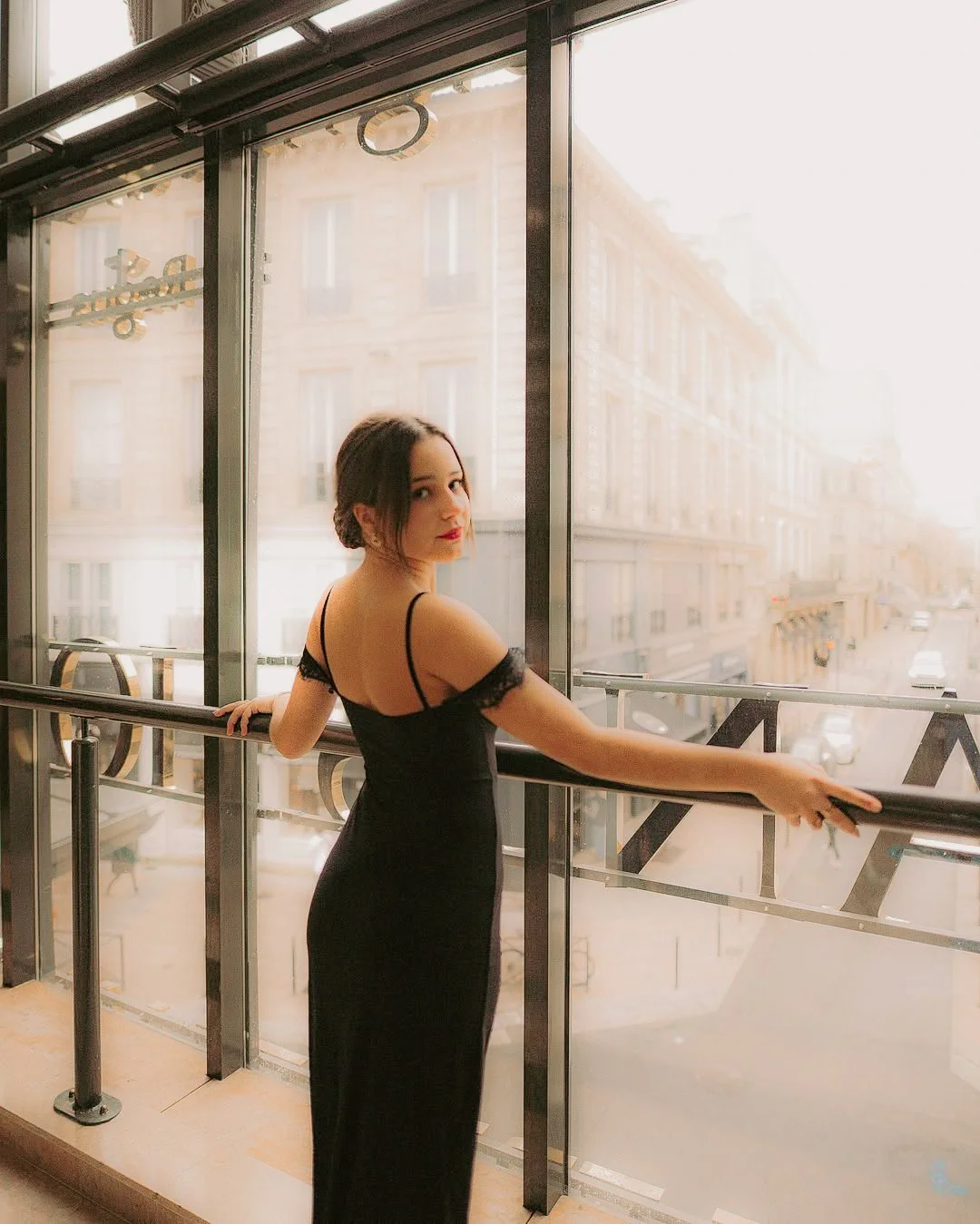 Jeune femme en robe noire, debout à une rampe en verre, regardant par une grande fenêtre d'un bâtiment, avec une vue sur une rue urbaine