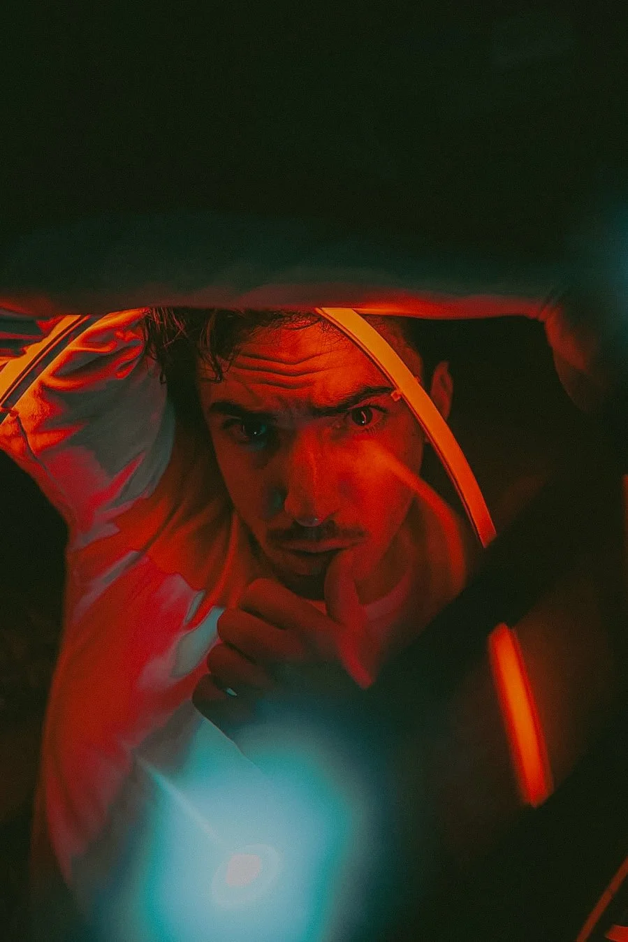 Un homme regarde vers le haut, éclairé par des lumières rouges et bleues, avec une expression sérieuse, portant une veste avec des bandes réfléchissantes.