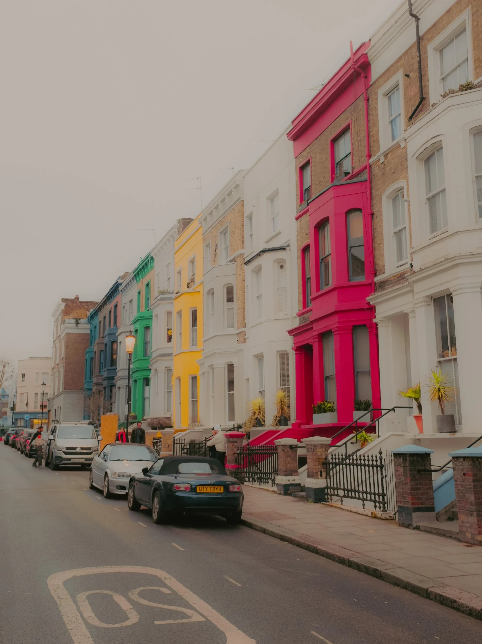 Immeubles colorés en face d'une rue avec des voitures garées et des piétons, dans une zone urbaine.