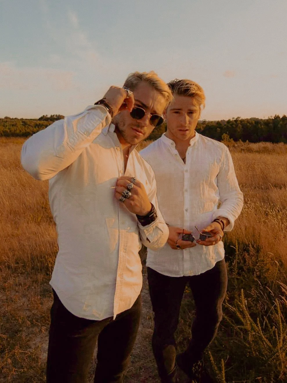 Deux jeunes hommes avec des cheveux blonds portant des chemises blanches, l'un avec des lunettes de soleil, dans un champ en plein air au coucher du soleil.
