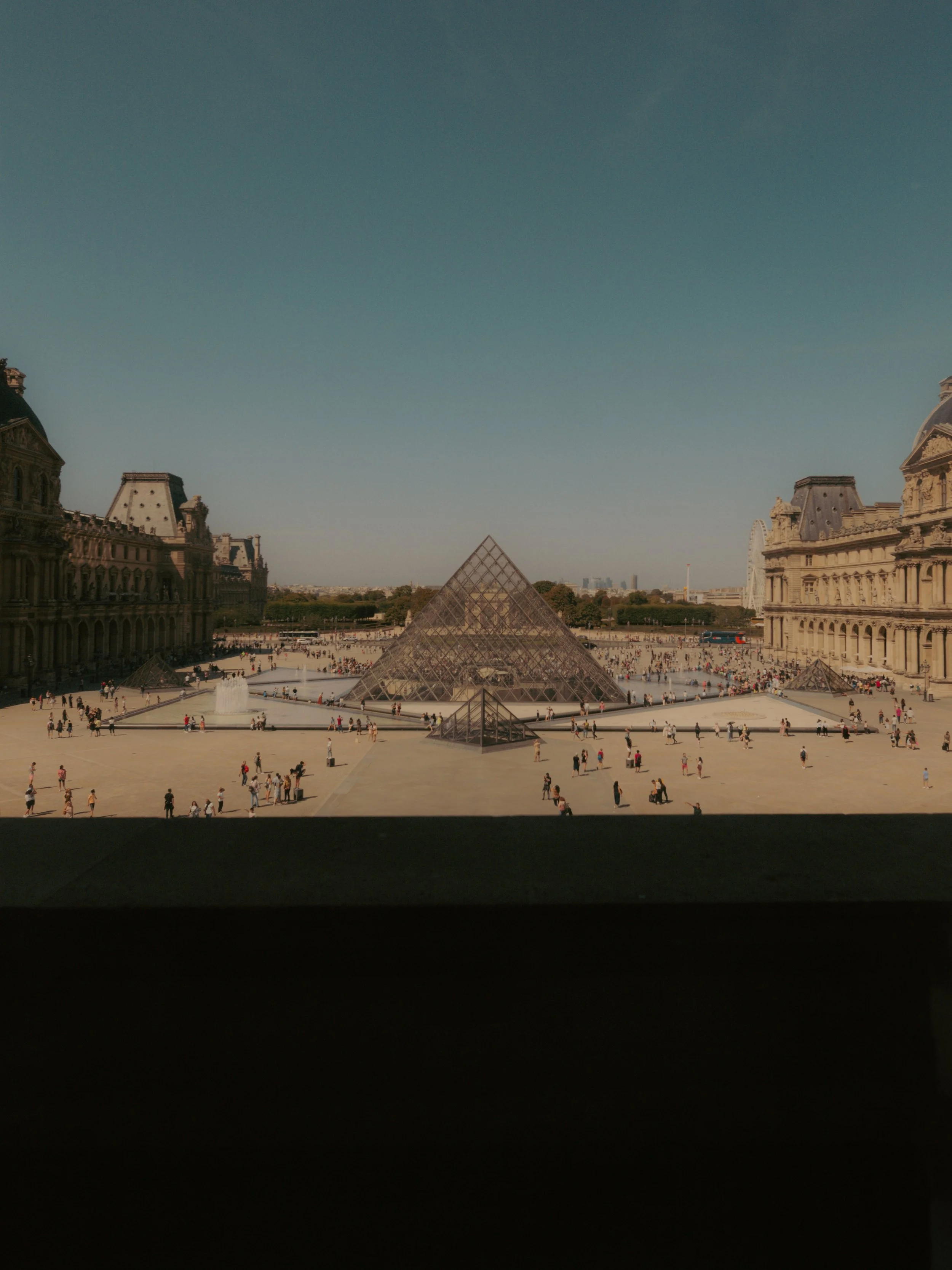 Vue du Louvre à Paris avec la pyramide en verre au centre, entourée de plusieurs bâtiments historiques, plusieurs touristes présents sur la cour.