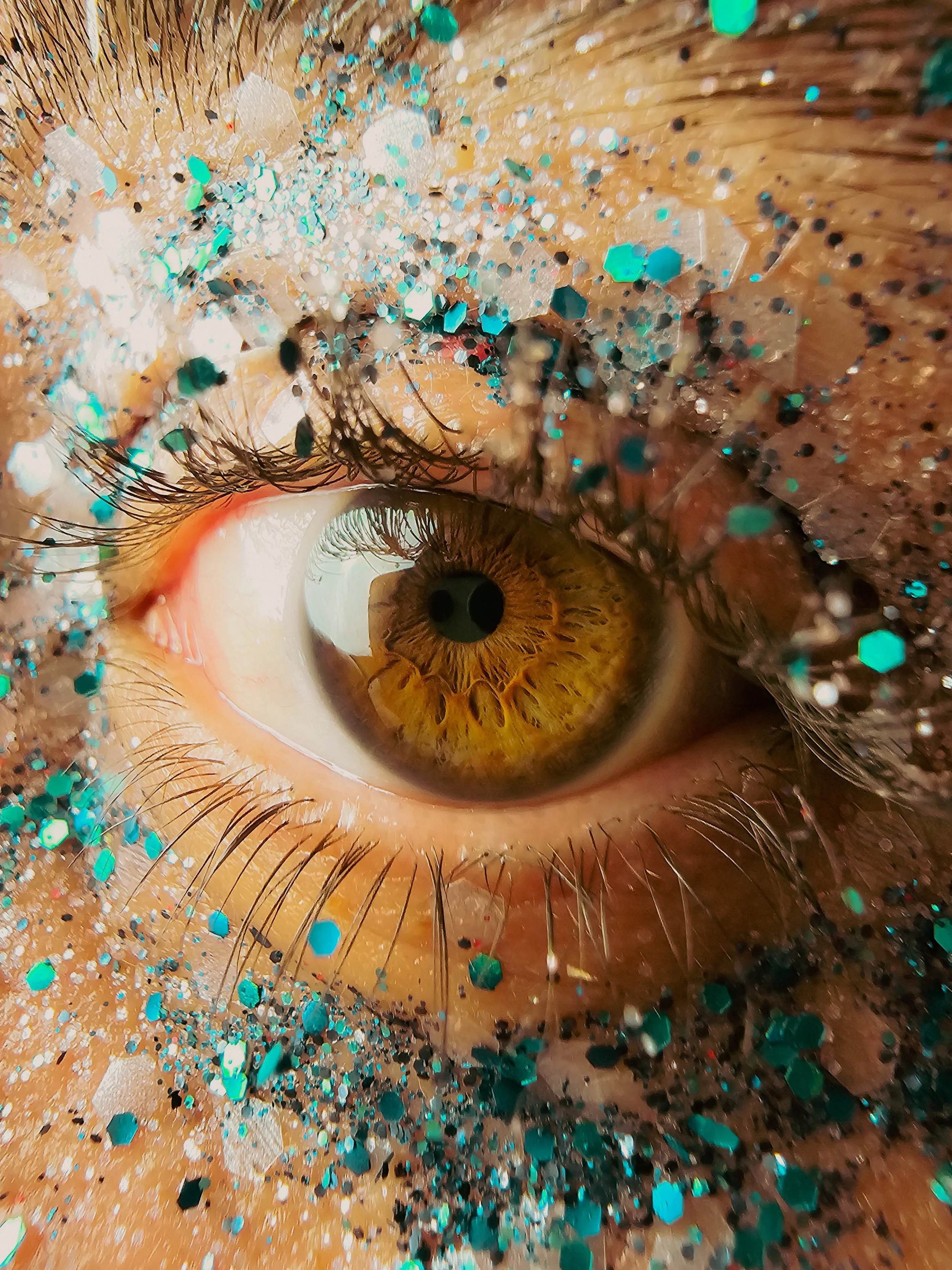 Un œil humain avec iris marron clair entouré de cils, partiellement recouvert de paillettes et de confettis bleus et argentés.