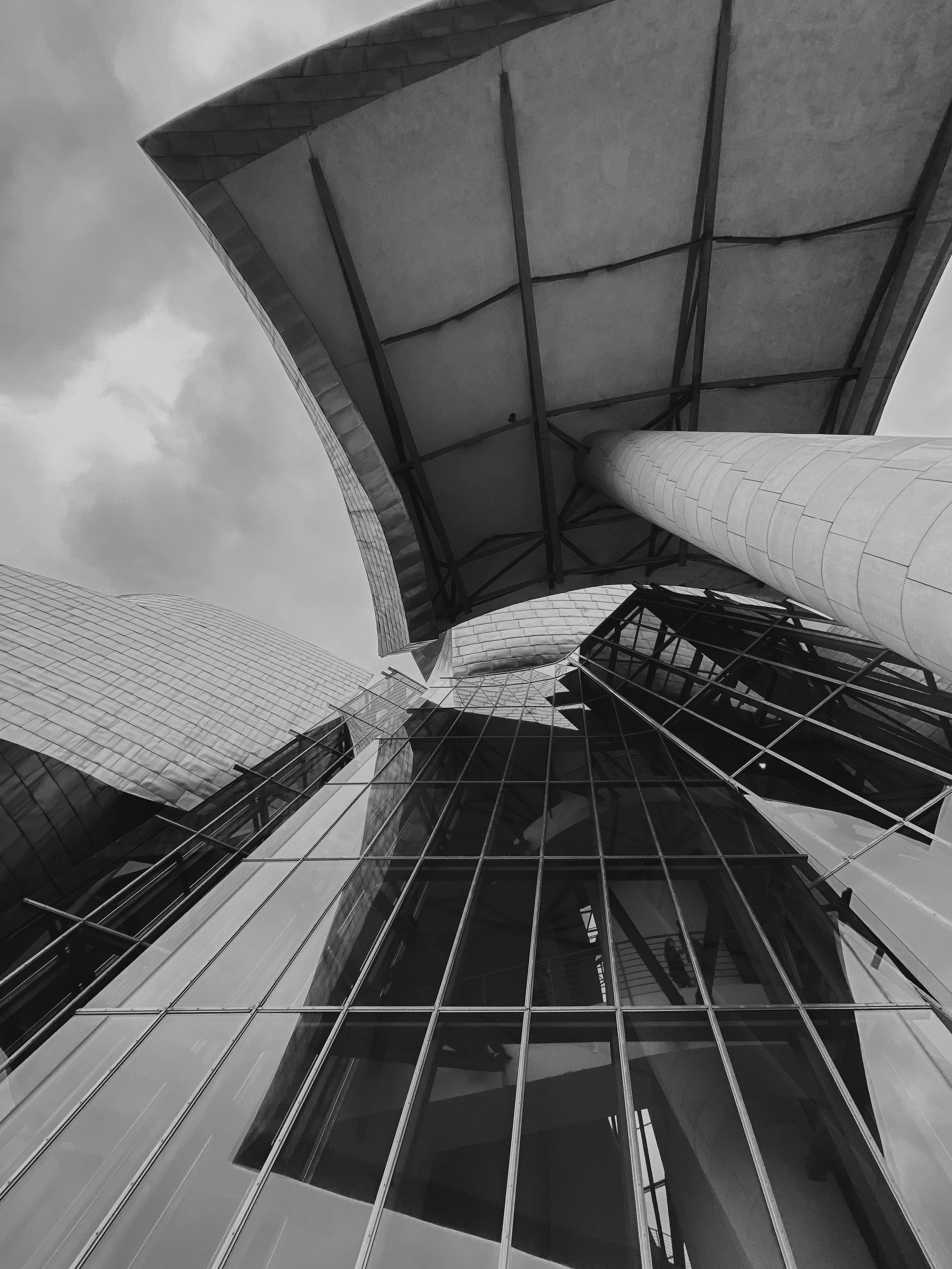 Vue en contre-plongée d'un bâtiment moderne avec des structures en verre et en métal, incluant une grande colonne et un toit incurvé, vue du sol vers le ciel avec des nuages.