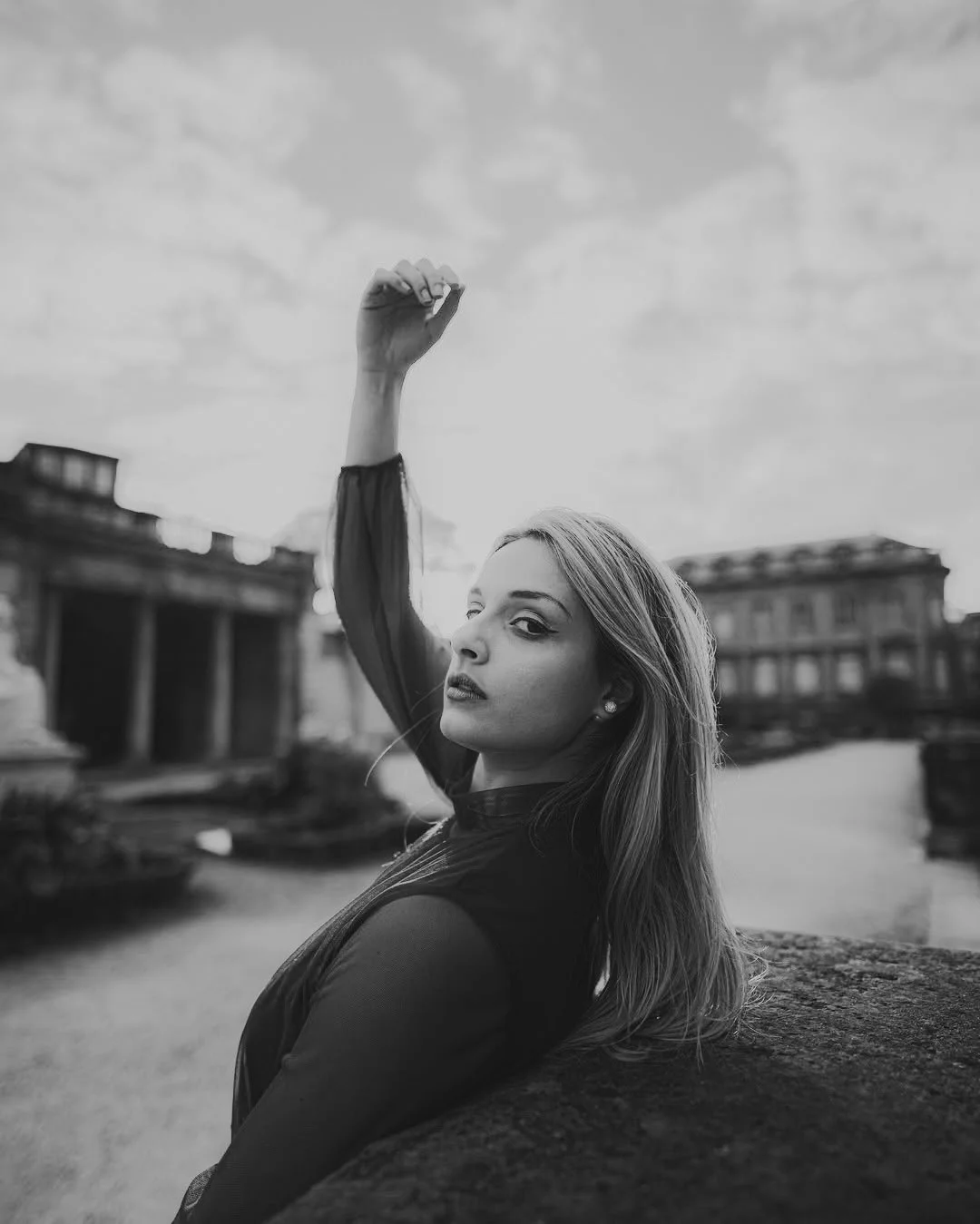Une femme pose sur un banc en extérieur avec des bâtiments anciens en arrière-plan, en noir et blanc.