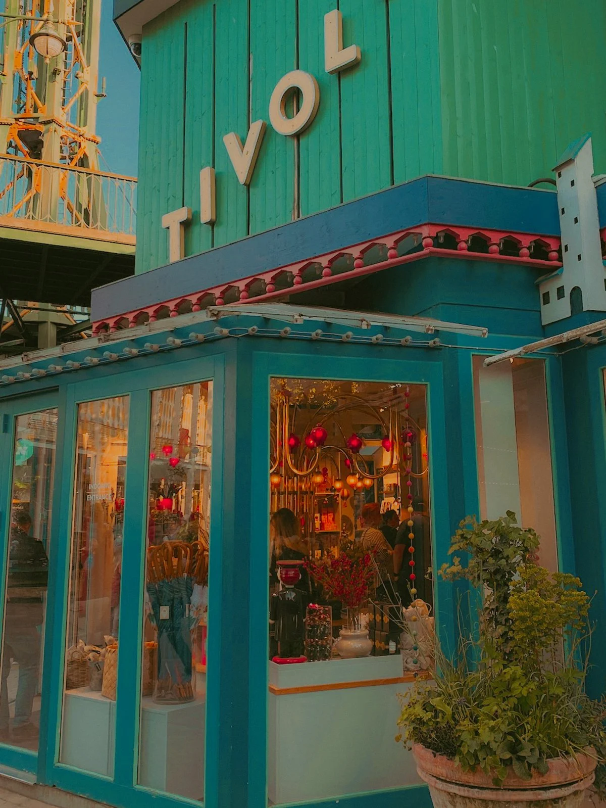 Façade d'un bâtiment coloré avec le nom 'TIVOLI' en lettres blanches. La structure est en bois turquoise avec de grandes fenêtres en verre montrant l'intérieur décoré avec des boules rouges suspendues. Il y a aussi un pot avec des plantes à l'extérie
