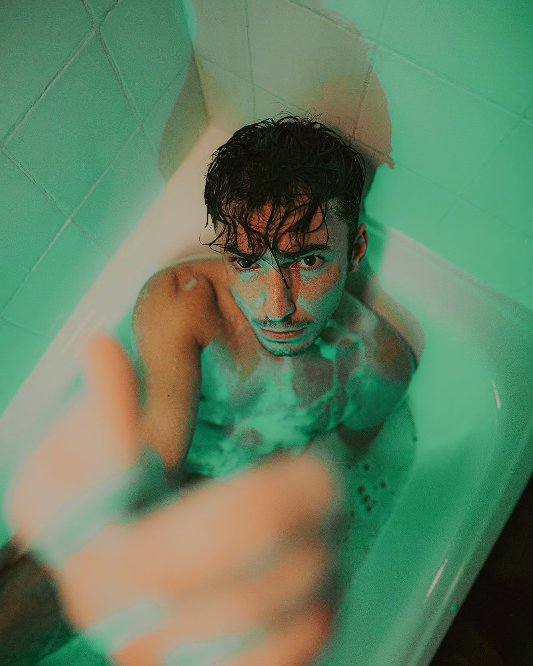 Un homme debout dans une baignoire, regardant vers le haut, avec de l'eau jusqu'à la poitrine, ses cheveux en désordre, dans une lumière verte.