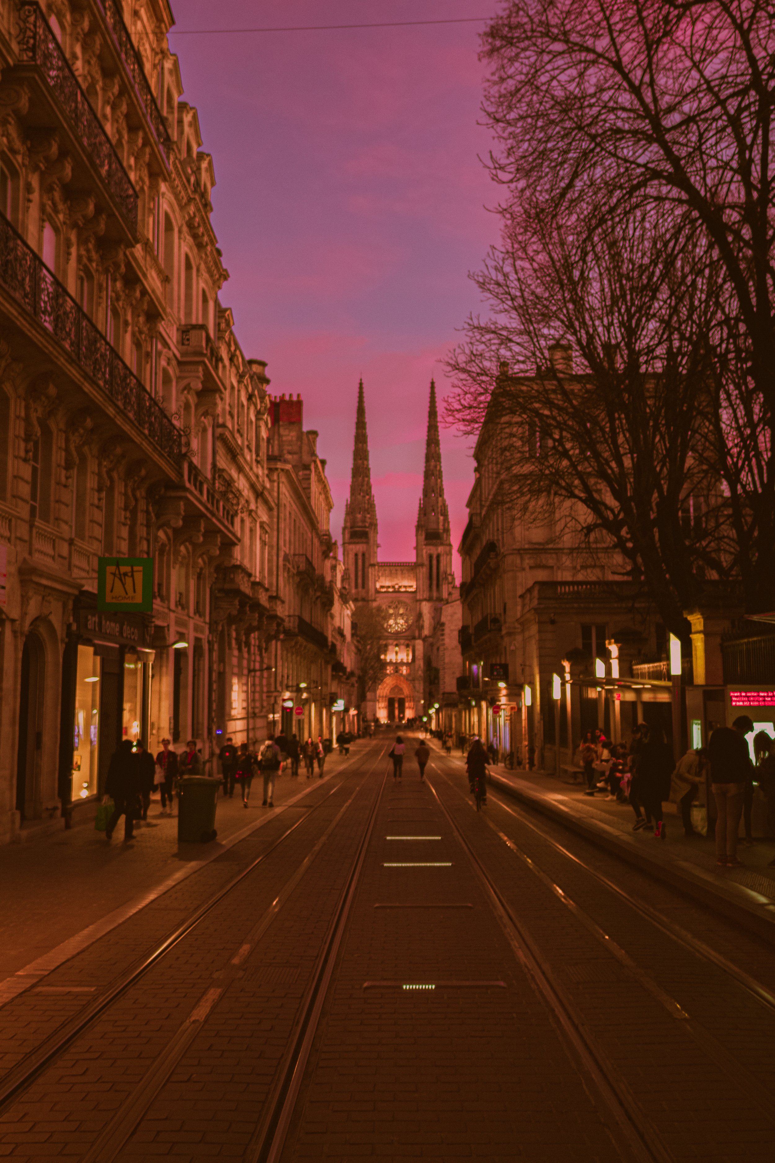 Une rue urbaine bordée de bâtiments historiques, avec une église gothique en arrière-plan, à l'aube ou au crépuscule, avec un ciel violet et rose.