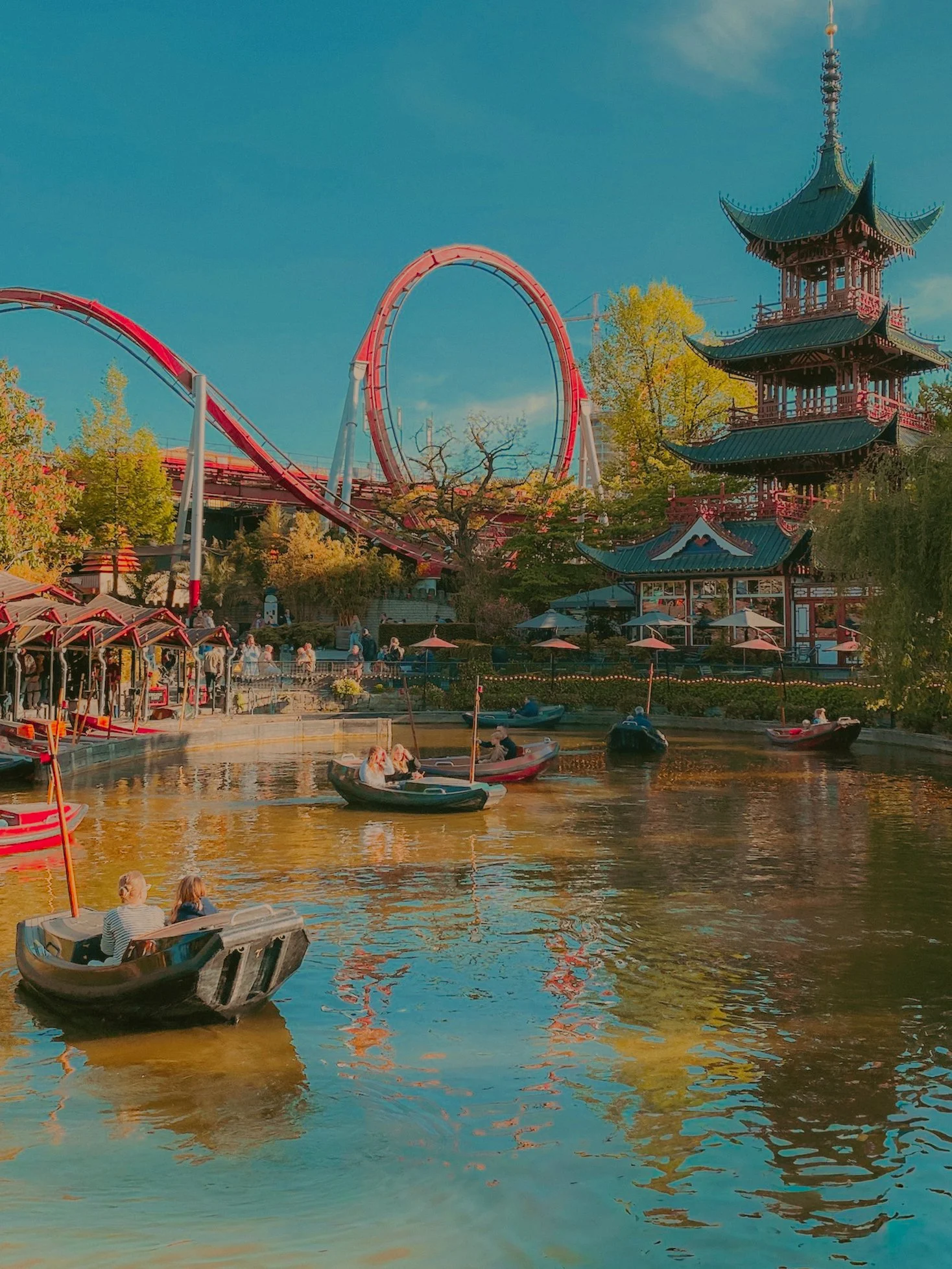 Une scène de parc d'attractions avec un lac où plusieurs bateaux à rames naviguent. En arrière-plan, on voit une grande roue rouge, une tour de manège à plusieurs étages de style asiatique avec un toit en pagode, et des arbres aux feuilles d'automne.