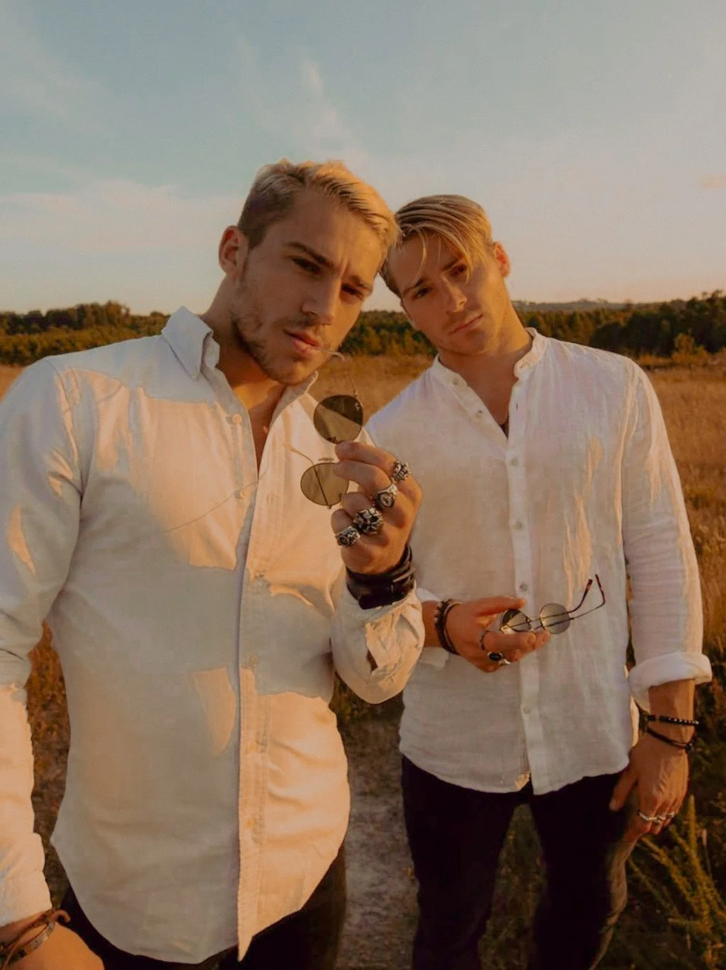 Deux jeunes hommes en chemises blanches posent dans un champ au coucher du soleil, portant des bijoux et des lunettes de soleil.