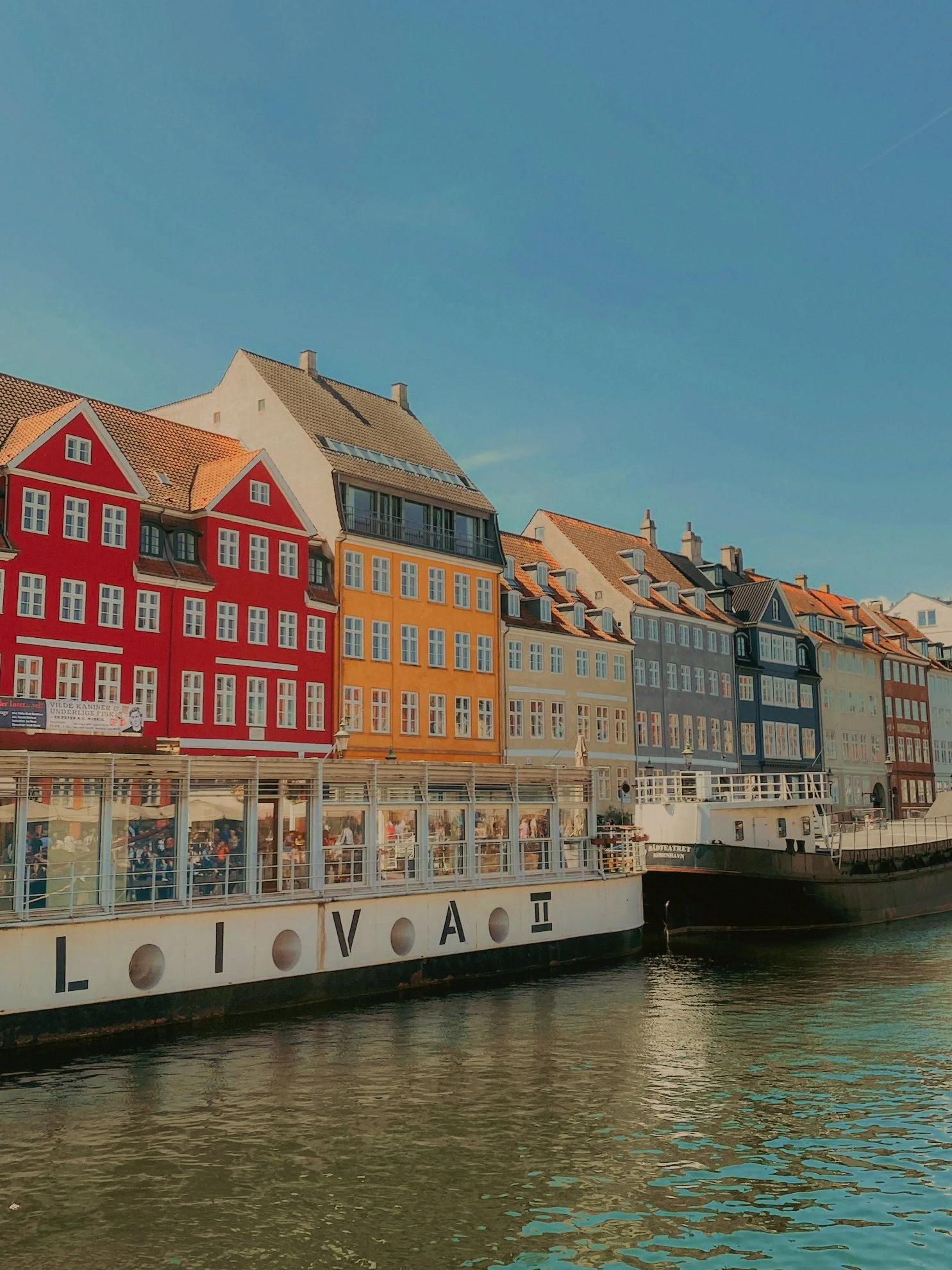 Maisons colorées alignées le long d'un fleuve, avec un bateau au premier plan et un ciel clair en arrière-plan.