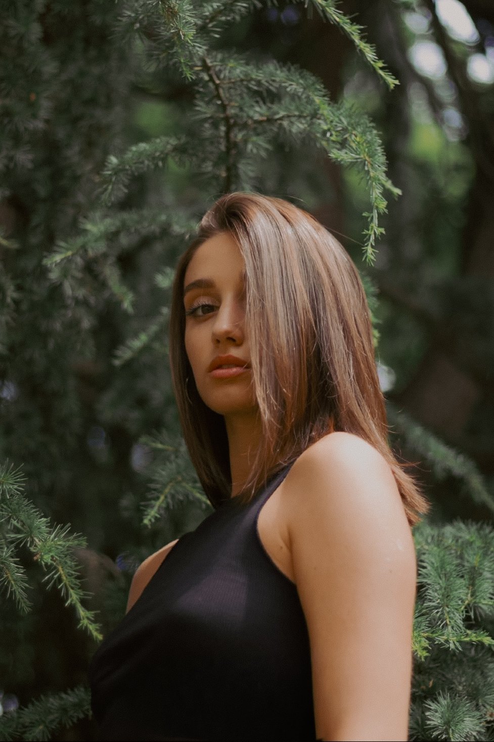 Une femme avec les cheveux bruns longs et droits, portant une robe noire, se tient devant un arbre vert feuillu.