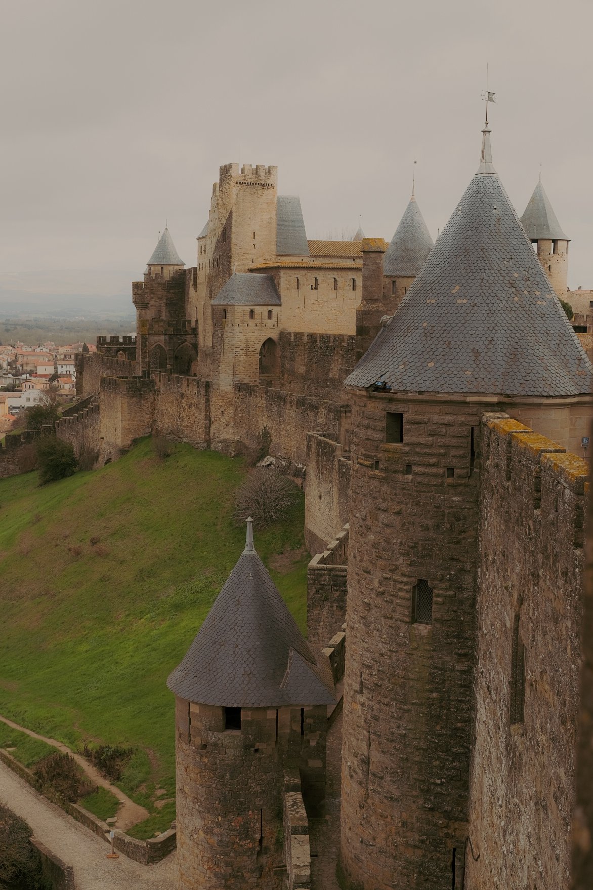 Un château médiéval avec des tours en pierre, situé sur une colline, entouré de murs fortifiés, en partie sous un ciel nuageux.