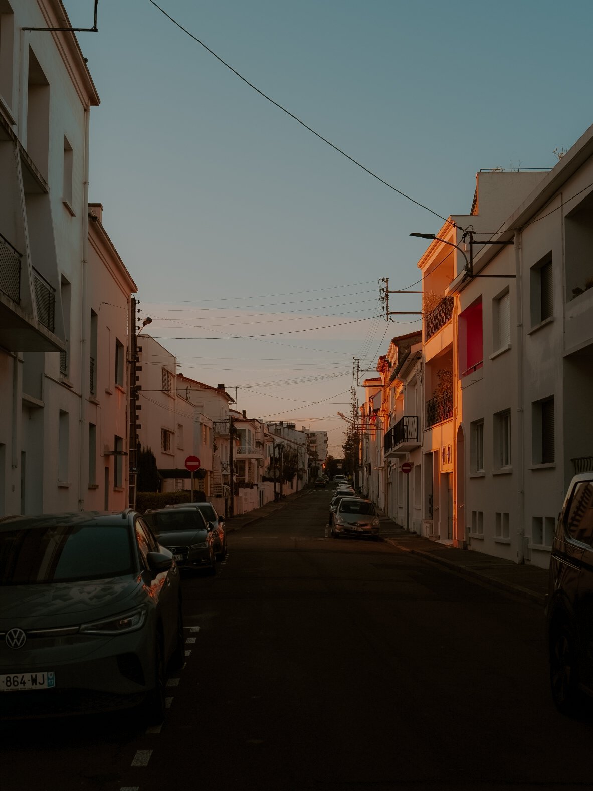 Rue résidentielle bordée d'immeubles blancs au coucher du soleil, avec voitures garées de chaque côté et câbles électriques dans le ciel.