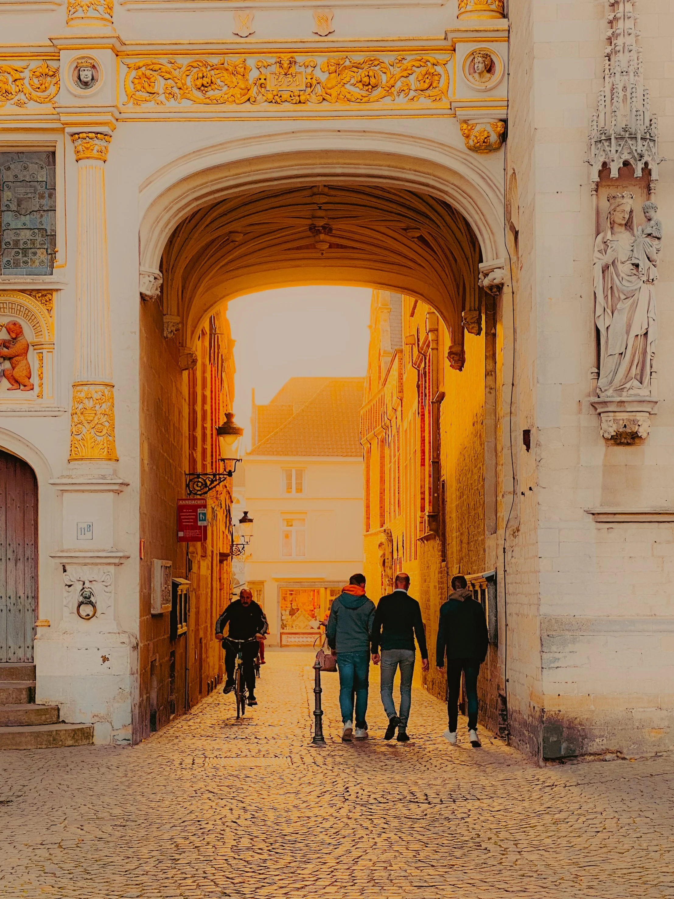 Vue d'une ruelle pavée médiévale avec un archeau en pierre ornée de sculptures et statues, illuminée par la lumière du coucher de soleil, avec quelques personnes et un cycliste passant.