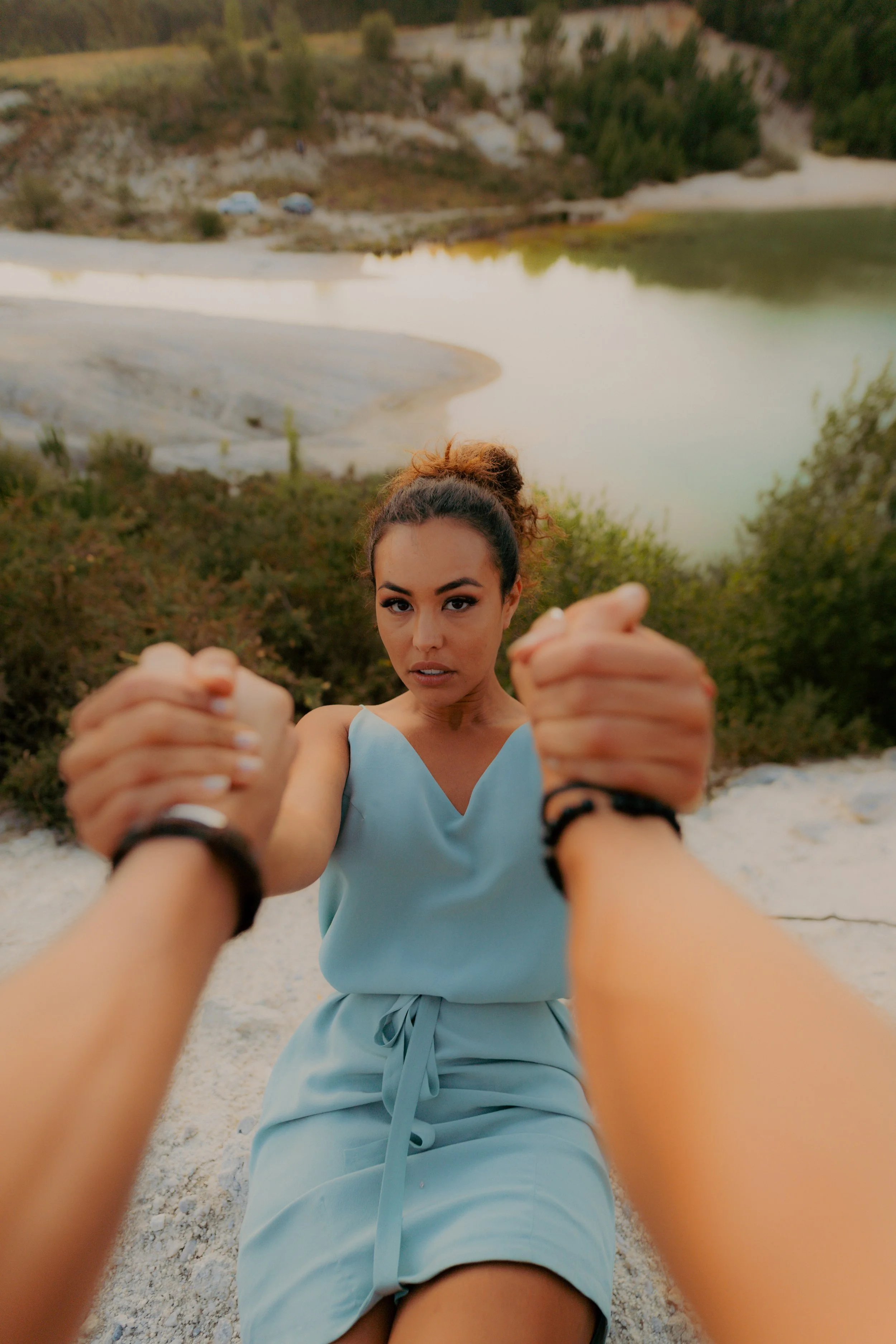 Une femme en robe bleue regarde l'objectif, tandis que deux autres personnes lui tiennent les mains en avant. En arrière-plan, on voit un lac entouré de collines et de végétation.