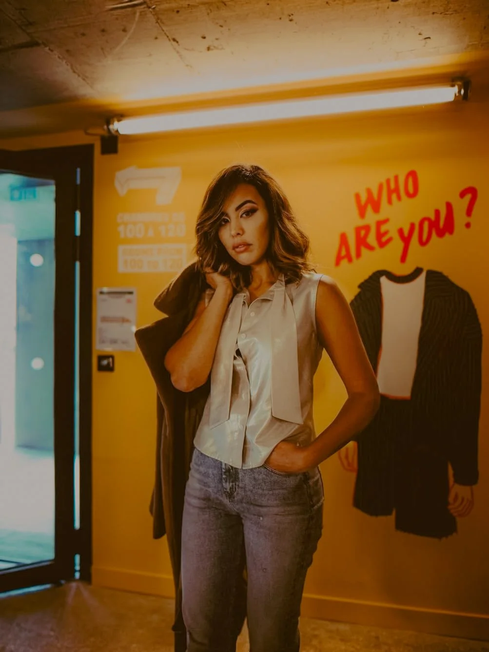 Jeune femme aux cheveux bouclés, portant un haut gris sans manches, pose dans un lieu avec un mur jaune décoré d'un dessin de veste et de la question 'Who are you?' en rouge.