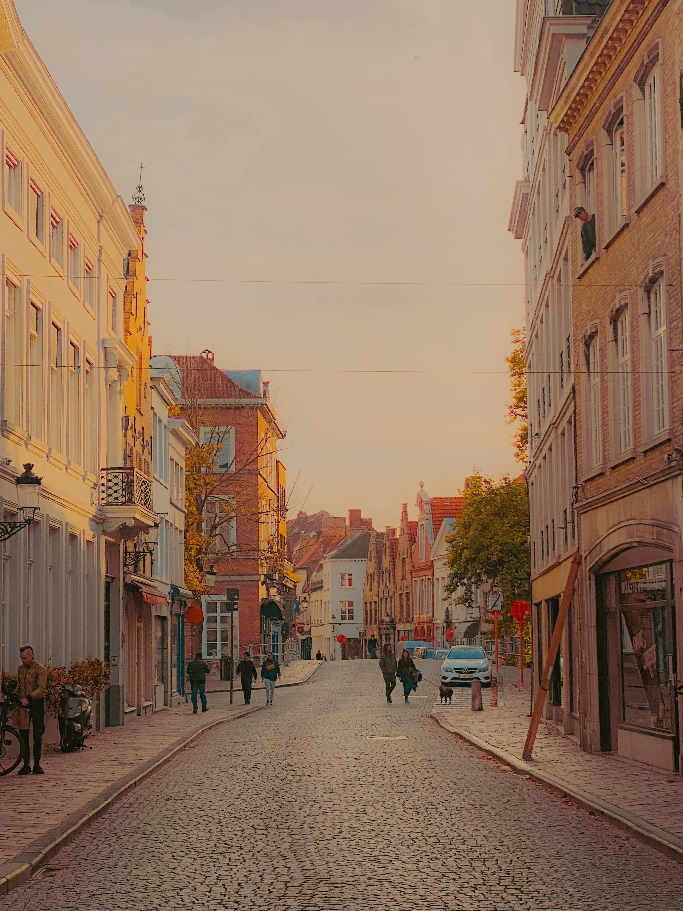 Une rue pavée dans une ville européenne avec des bâtiments anciens, des personnes marchant, et quelques voitures stationnées, sous un ciel au coucher du soleil.