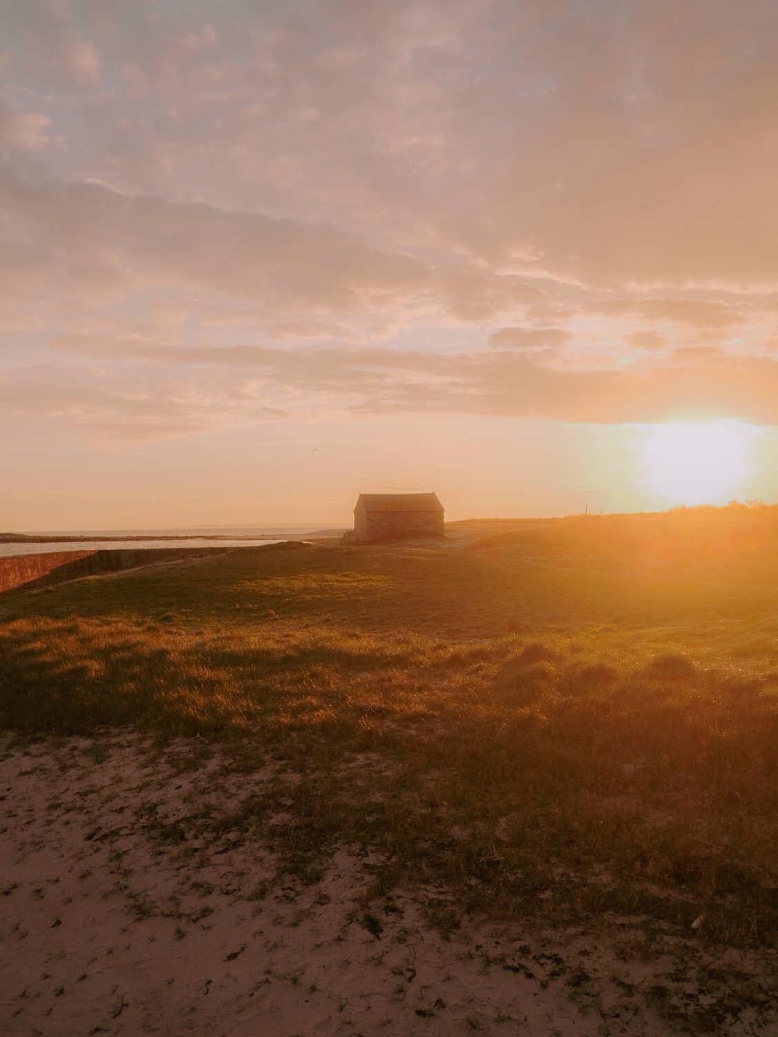 Coucher de soleil sur un paysage avec une maison en bois, de l'herbe et du sable, dans un environnement tranquille.