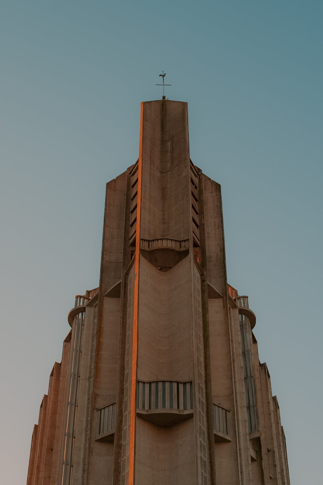 Photo d'un bâtiment en béton avec des balcons arrondis et des fenêtres verticales, sommet avec une girouette en forme d'oiseau sur une planche à voile, contre un ciel clair.
