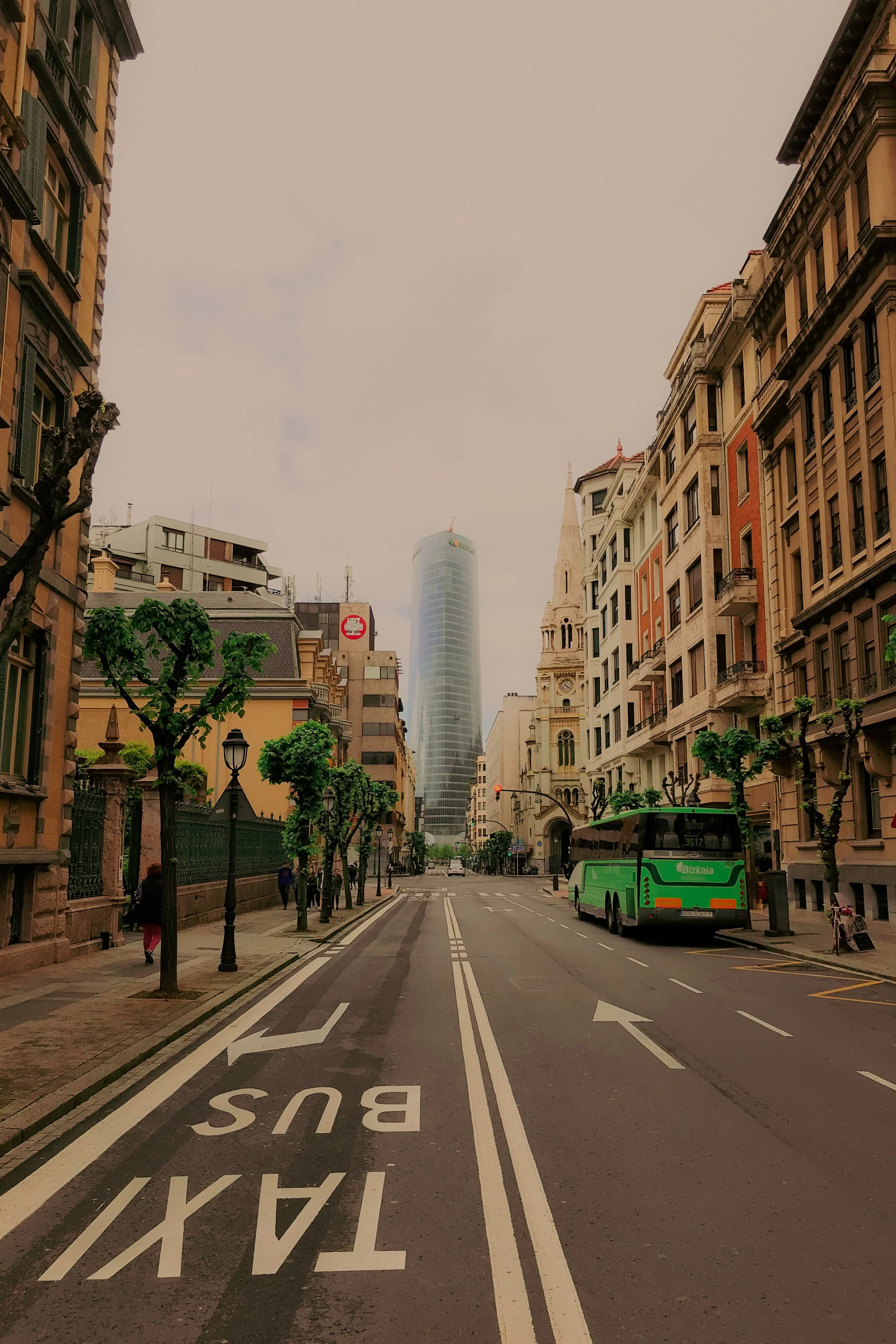 Rue urbaine avec des bâtiments historiques et modernes, bus vert stationné sur le côté, arbres taillés, ciel nuageux