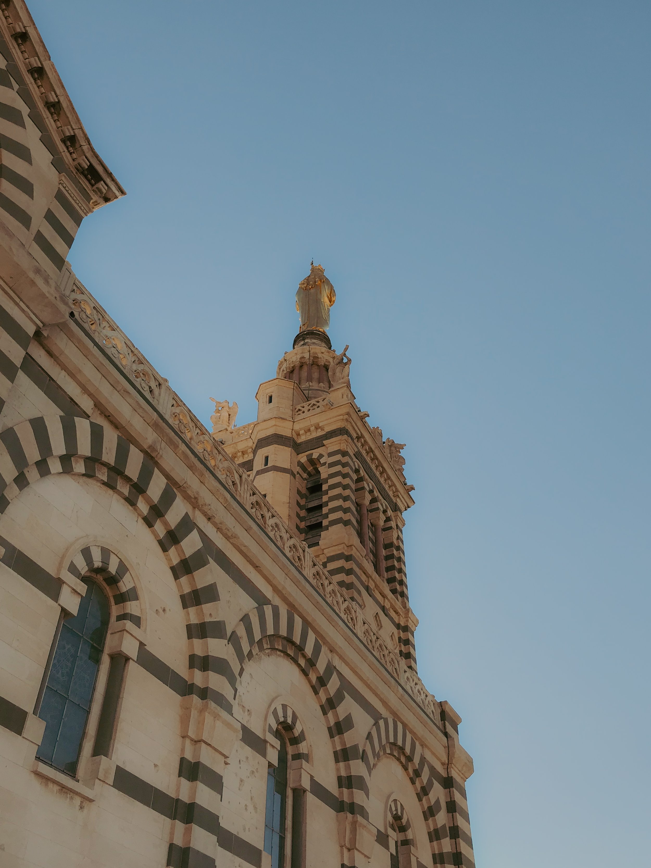 Vue d'une église avec des arches en pierre blanches et noires, une tour avec des fenêtres, un toit en forme de cloche, et une statue dorée en haut, sous un ciel dégagé.
