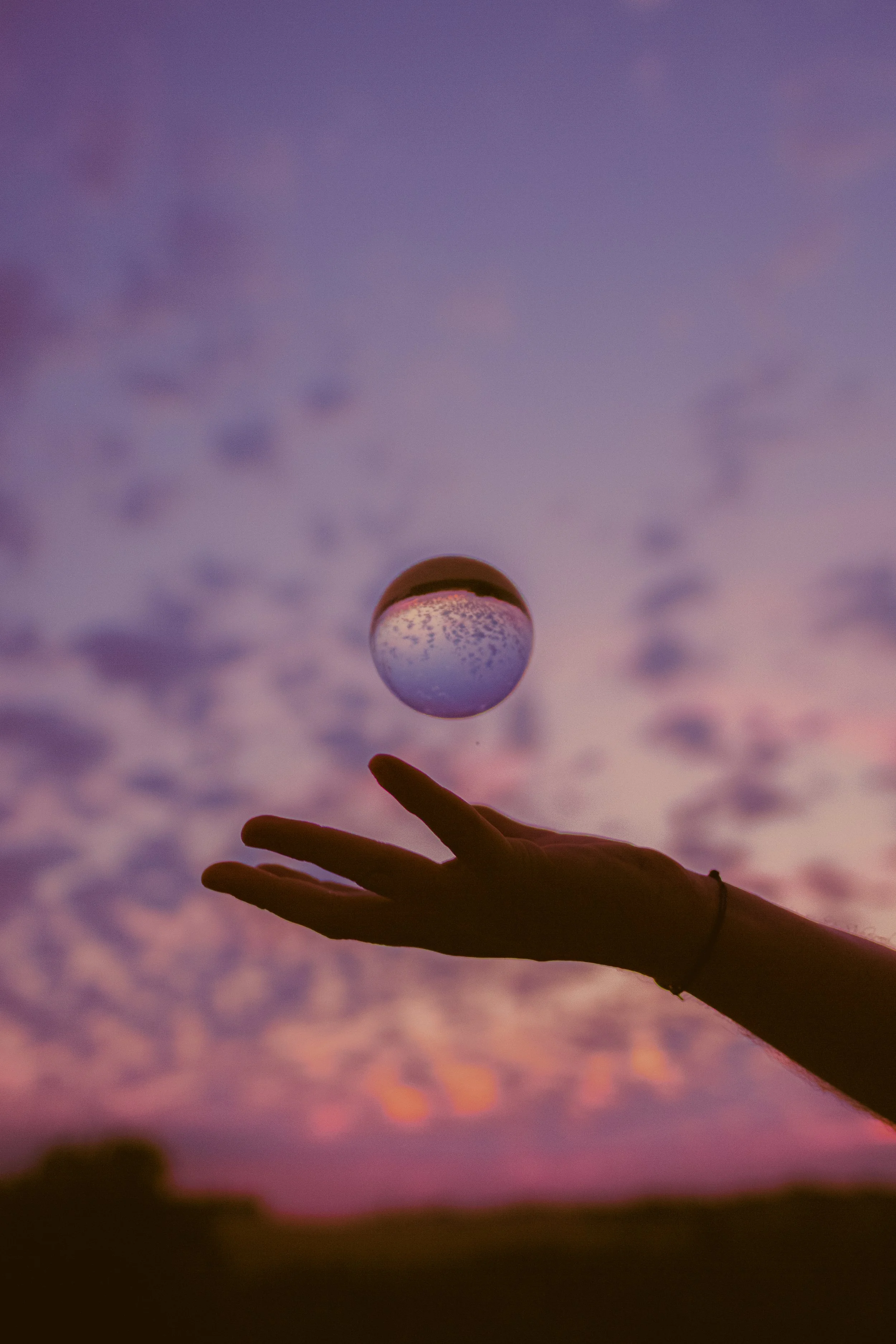 Une main silhouette tenant un boule de cristal contre un ciel au coucher du soleil avec des nuages violets et roses.